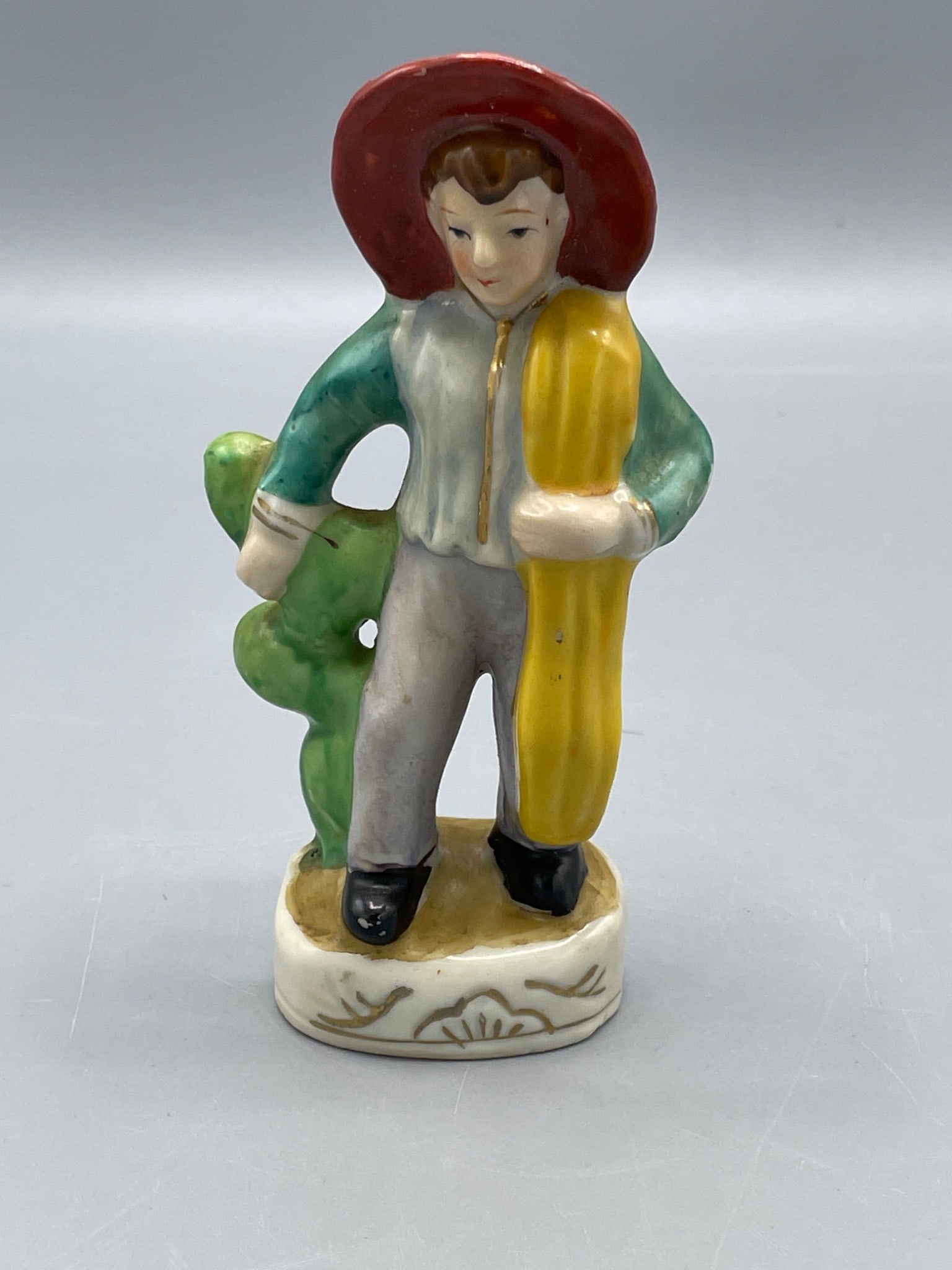 Hand-Painted Figurine from Japan, Approximately 5 Inches Tall: This is a ceramic figurine hand-painted in Japan. It stands approximately 5 inches tall and depicts a figure wearing a hat and carrying an object. The piece has a hollow base with the marking
