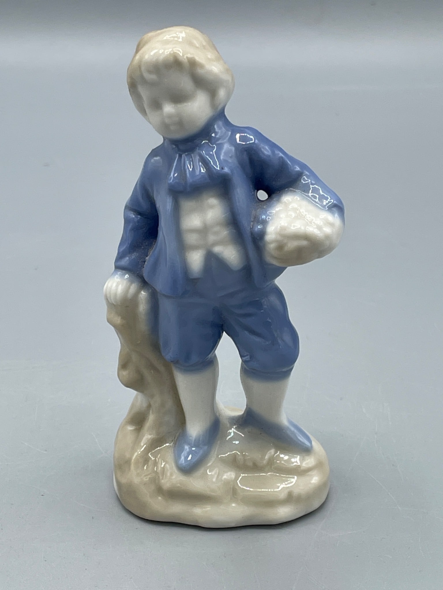 Porcelain Figurine of a Boy with 'FOREIGN' Marking, Approx. 5 Inches: This listing is for a small porcelain figurine measuring approximately 5 inches in height. The figurine depicts a young boy holding an object, dressed in period-style clothing and painted in blue and 