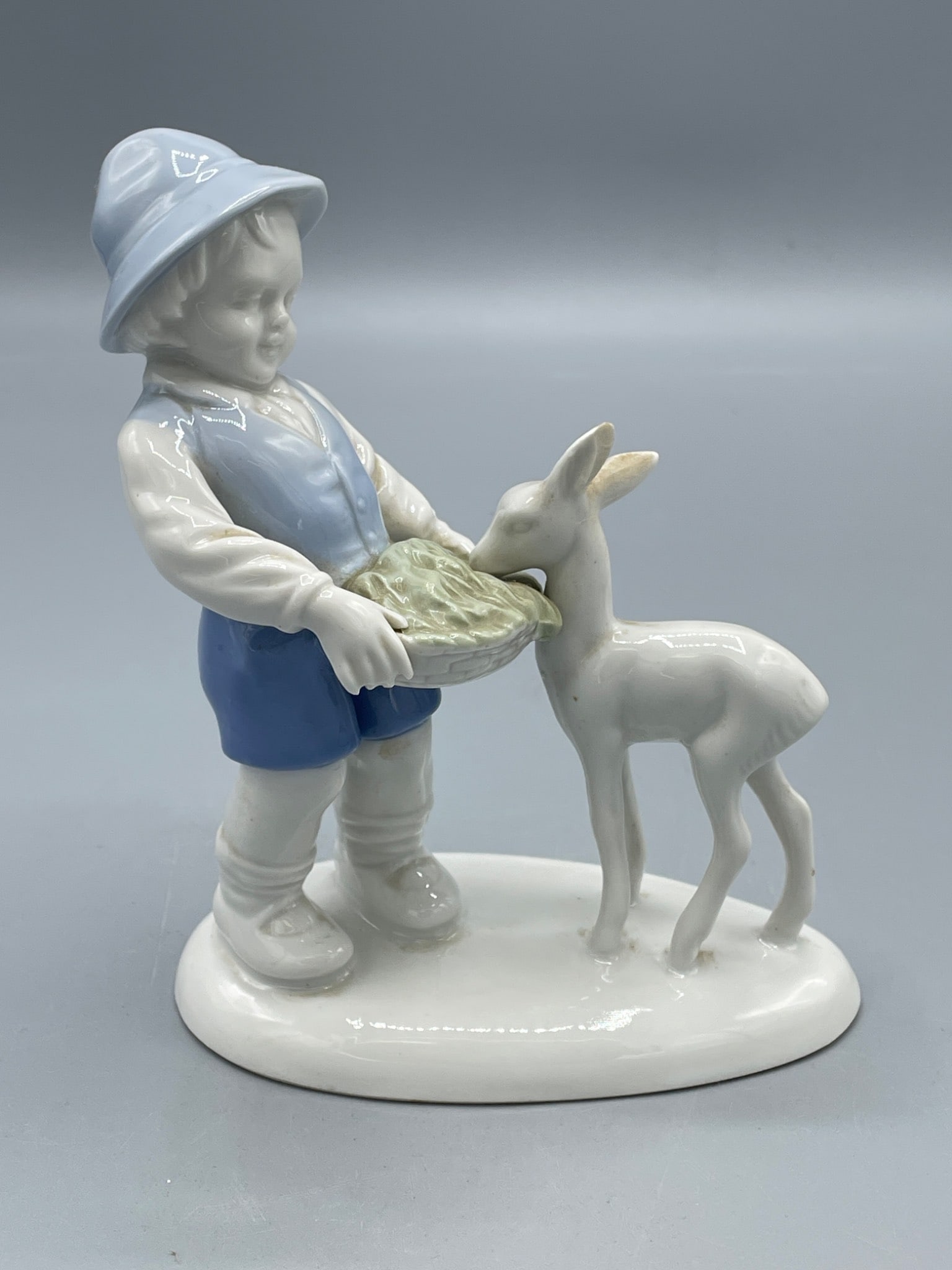 Porcelain Figurine of Boy Feeding Deer, Approx. 6 inches: Porcelain figurine depicting a boy wearing a hat and vest feeding a deer. The figurine is painted in shades of blue, white, and light green. The approximate height is 6 inches as shown in the image.