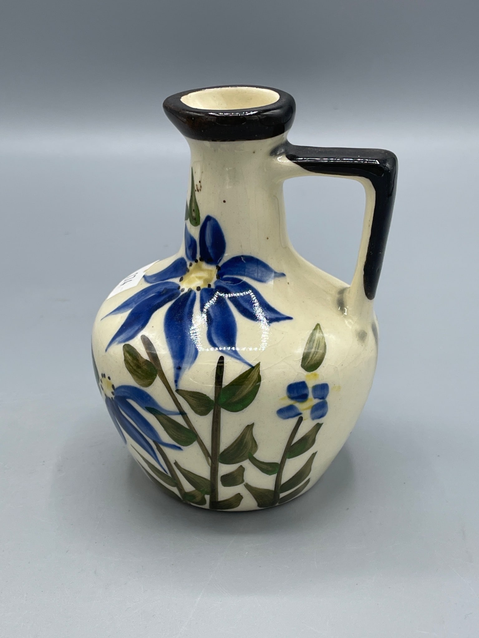 Decorative Ceramic Jug With Hand-Painted Floral Design: Small ceramic jug with a hand-painted floral design in blue and green on a cream background. Features a distinctive open cutout design on one side and a black-painted handle and rim. Approximate heigh