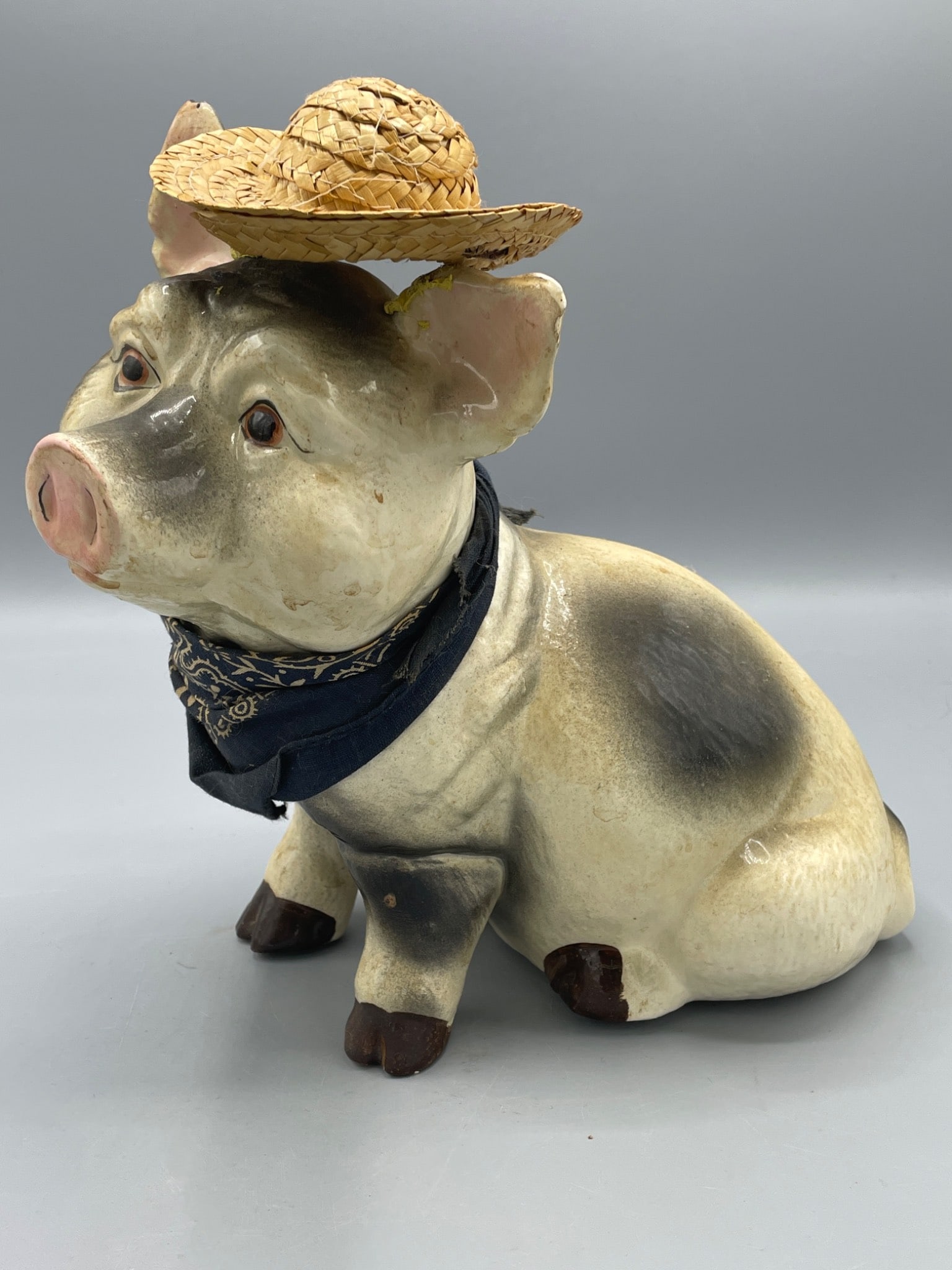 Ceramic Pig Figurine with Straw Hat and Bandana: A ceramic pig figurine featuring black and white coloration, wearing a blue bandana and a removable woven straw hat. Hollow construction with an open bottom; measures approximately 9 inches tall, as