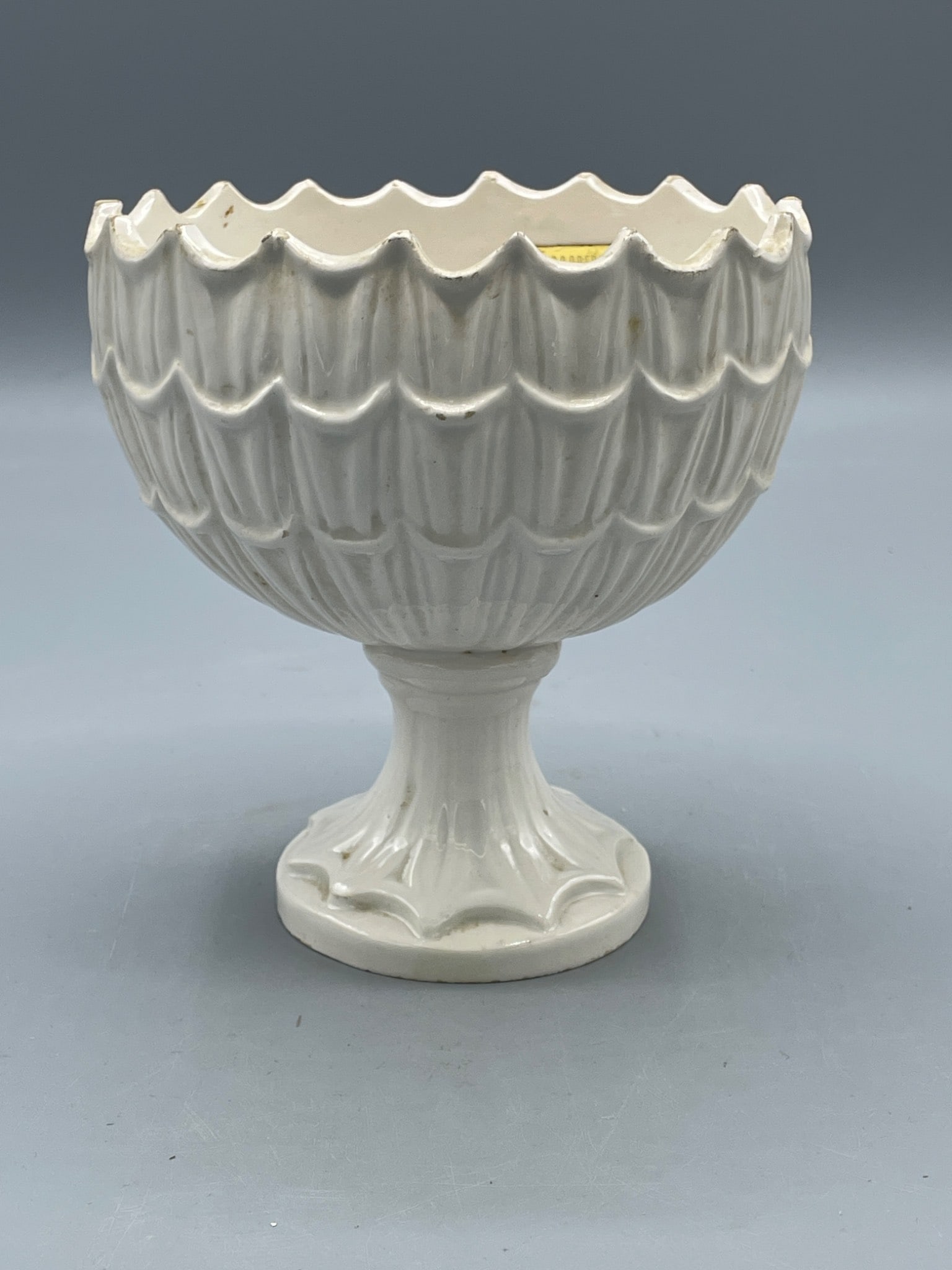 White Ceramic Pedestal Bowl by C.S. Goodfriend Imports: A white ceramic pedestal bowl featuring a scalloped edge and textured design, standing approximately 6 inches high. Labeled on the interior with a gold sticker from C.S. Goodfriend Imports, Inc. The