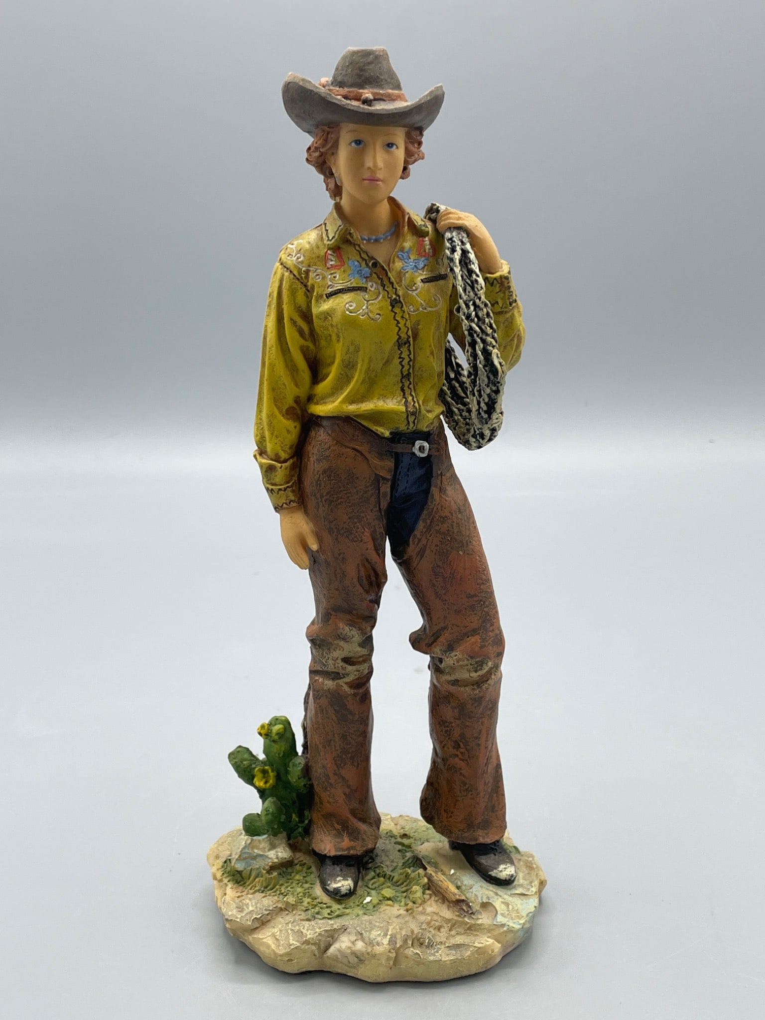 Cowgirl Figurine With Hat, Lasso, and Cactus Detail: This figurine depicts a cowgirl dressed in a yellow shirt with embroidered details, brown chaps, and a cowboy hat, holding a lasso. The figure stands on a textured base with cactus accents. Measuring