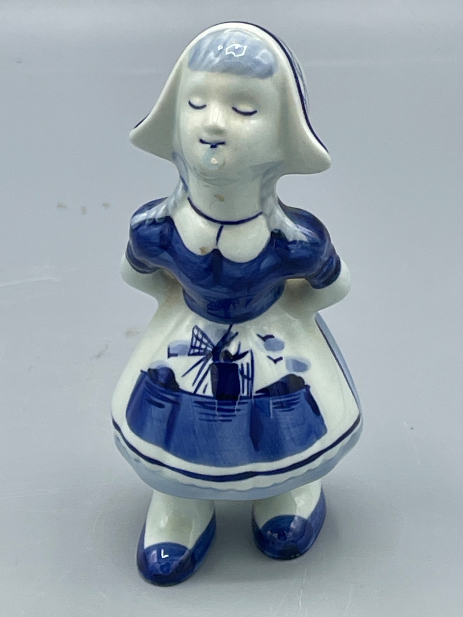 Hand-Painted Ceramic Dutch Girl Figurine with Windmill Design, Approx. 4.25 Inches Tall: A ceramic Dutch girl figurine hand-painted in blue and white, featuring windmill imagery on the front of her dress. The figure stands approximately 4.25 inches tall, as measured against a ruler. Small