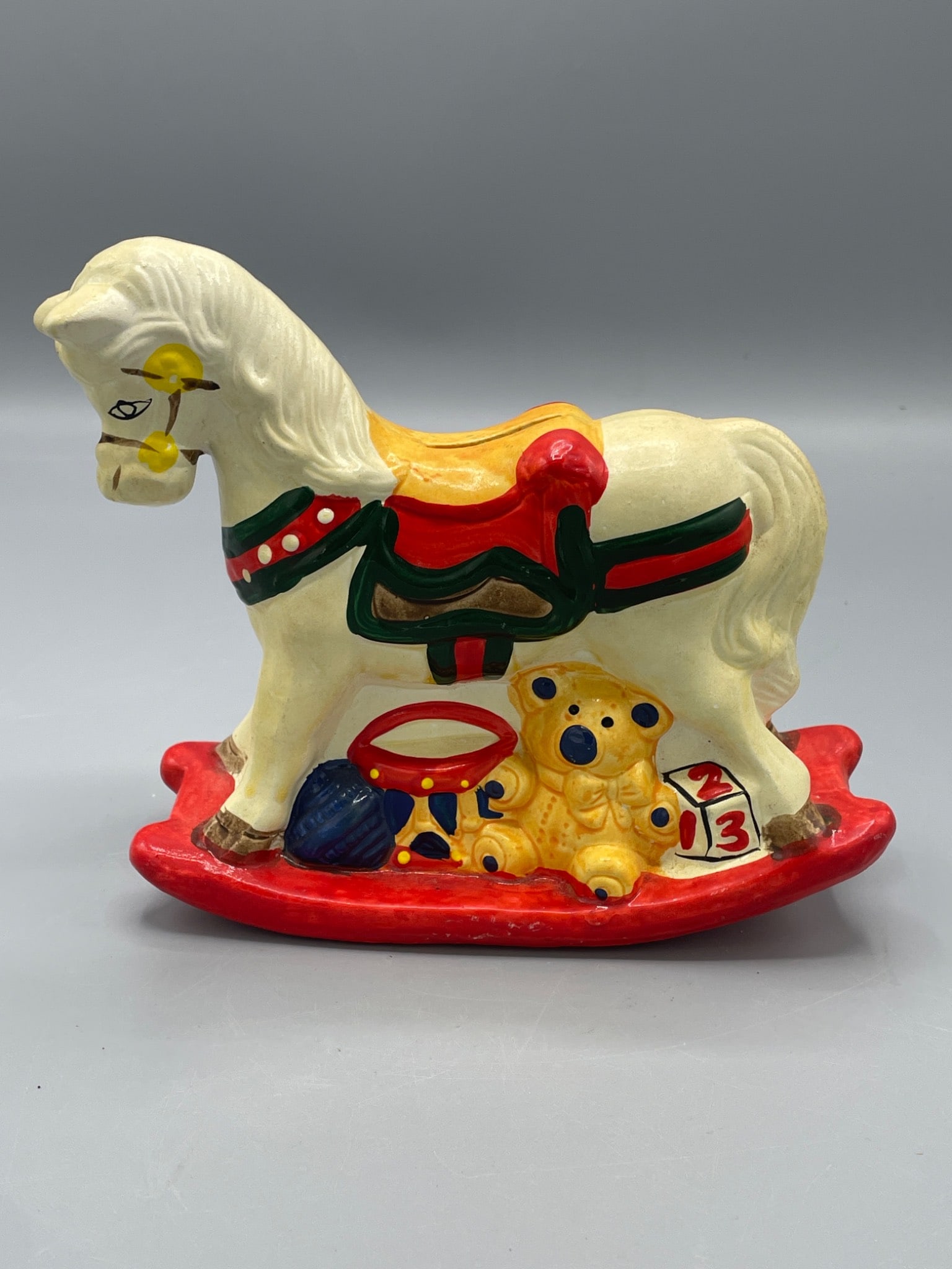 Painted Ceramic Rocking Horse Coin Bank with Slot and Rubber Stopper: A ceramic rocking horse coin bank featuring a painted design with a white horse, red saddle, and decorative accents. It includes a coin slot on the back and a rubber stopper on the base for access to