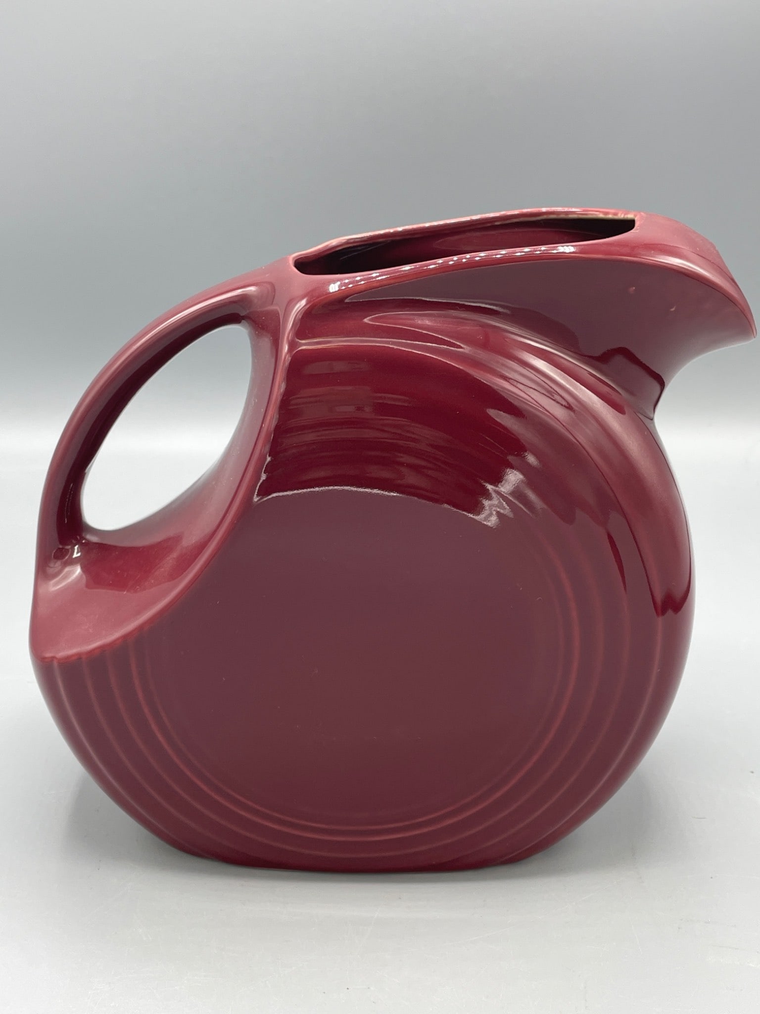 Red Ceramic Disc Pitcher with Rounded Handle and Art Deco Design fiesta brand: This red ceramic pitcher has a disc shape with a rounded handle and features an Art Deco design aesthetic. It measures approximately 8 inches in height as shown in the provided images. The base shows