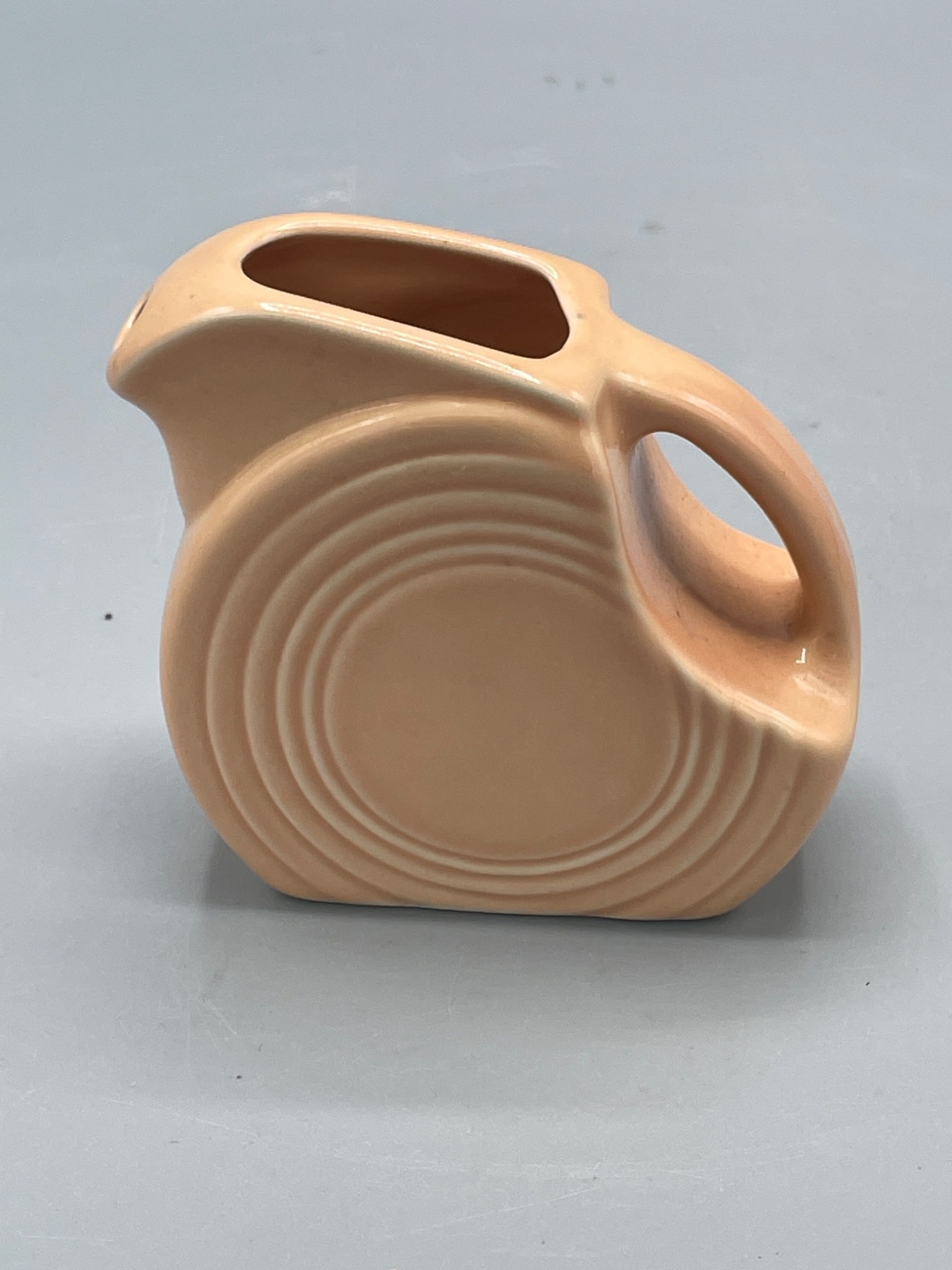 Fiesta Brand Ceramic Disc Pitcher in Peach Glaze, Approximately 6 Inches Tall: A ceramic disc pitcher by Fiesta brand featuring a peach glaze finish. The pitcher measures approximately 6 inches in height. The bottom is marked with the Fiesta logo. Condition appears consistent