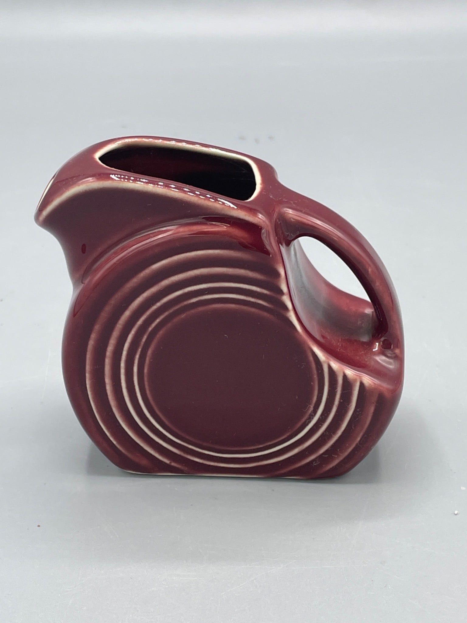 HLC Fiesta Mini Disc Pitcher in Maroon Glaze, Made in USA: This is a mini disc pitcher by Homer Laughlin China Company (HLC), part of the Fiesta line. The pitcher is finished in a maroon glaze and features the classic concentric ring design. The base is
