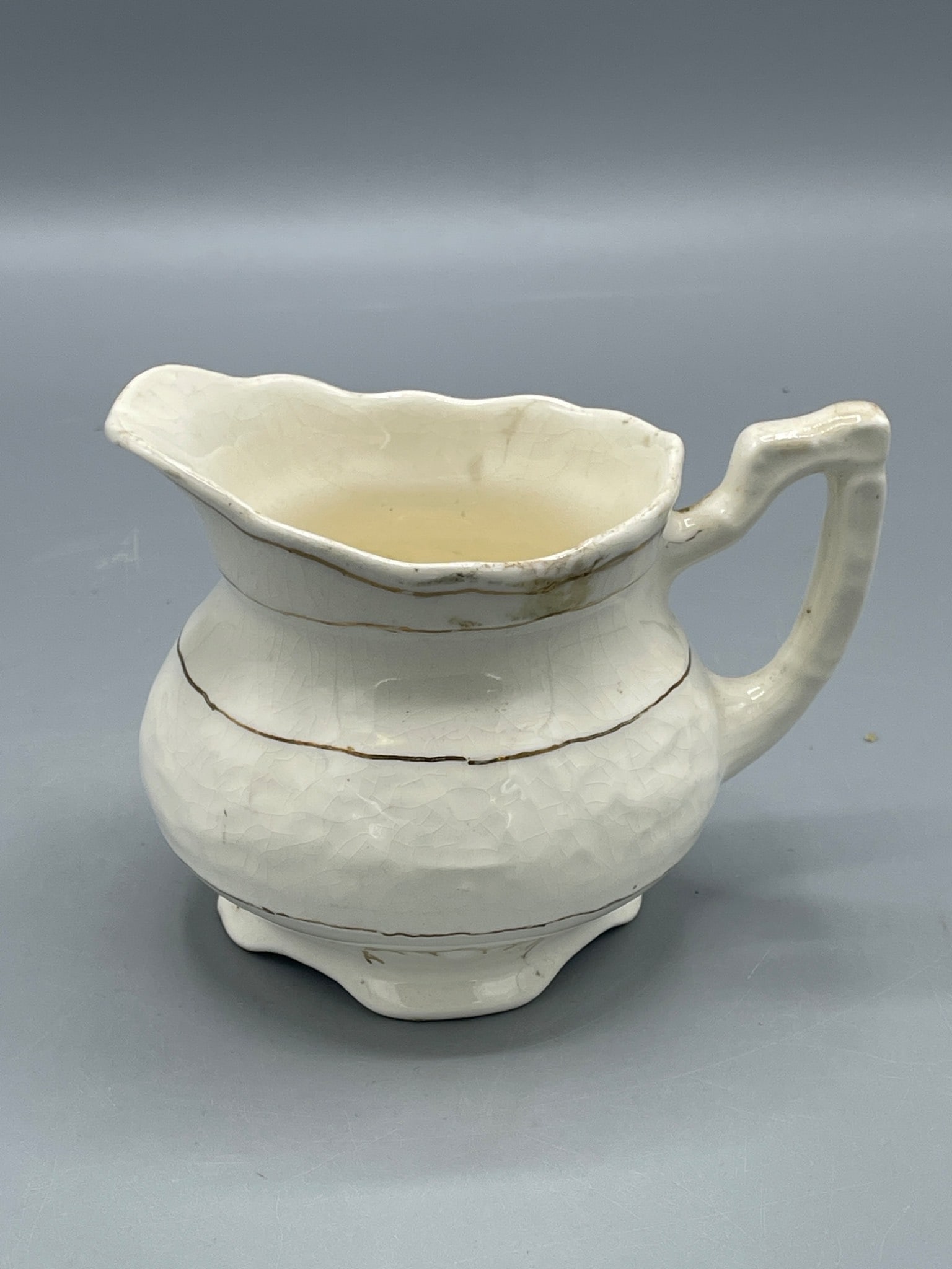 Cream-Colored Ceramic Pitcher with 'Hauso 1956' Inscription: Small ceramic pitcher featuring a cream-colored glaze with scalloped edges and a single handle. The base is inscribed with 'Hauso 1956.' The item shows signs of wear, including staining and minor