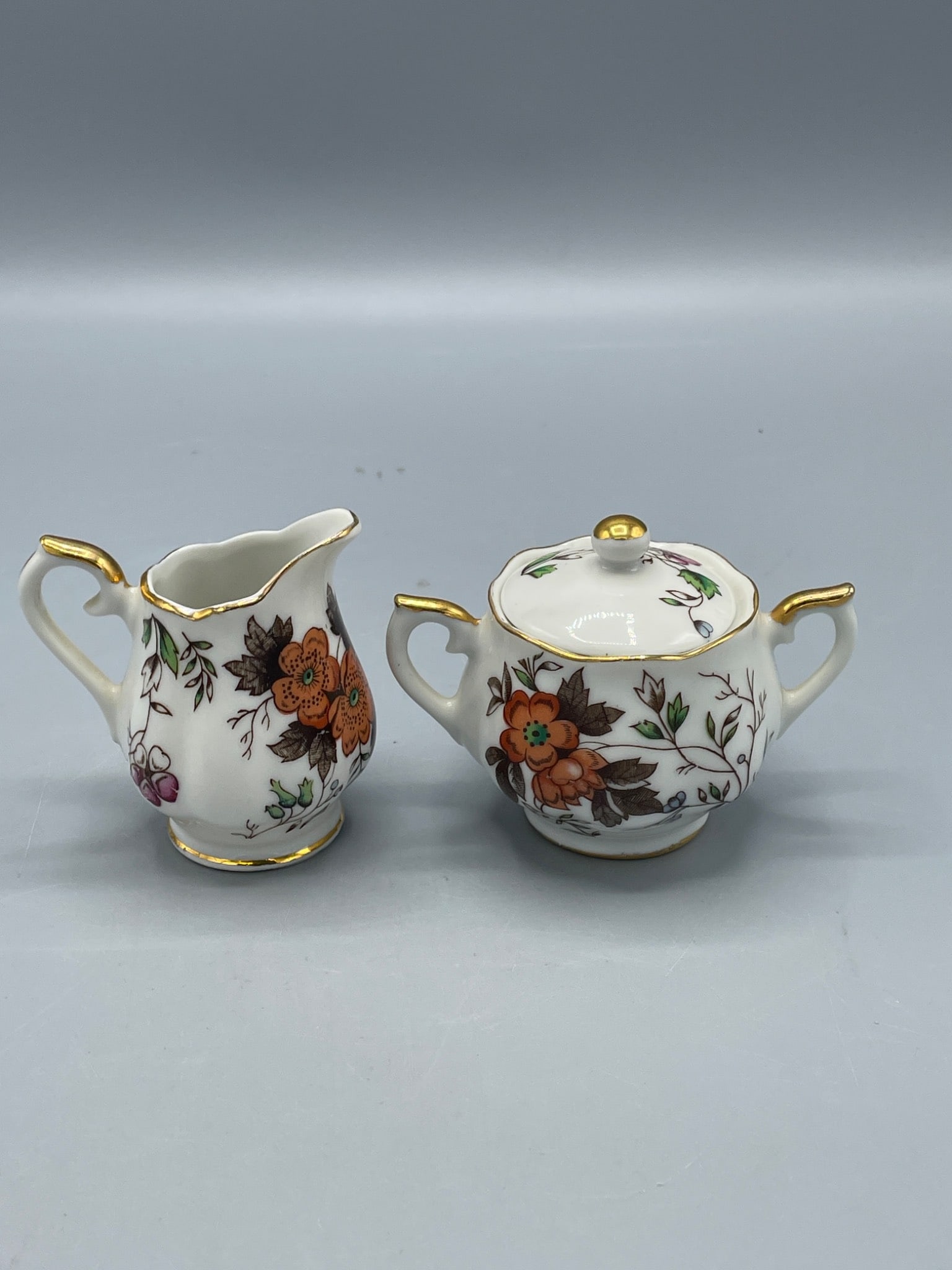 Enesco Japan Creamer and Sugar Bowl Set with Floral Design and Gold Accents: This lot features a matching Enesco Japan creamer and sugar bowl set, identified by 'Enesco Imports Japan' stickers on the base. The pieces showcase a floral design with vibrant orange, red, and