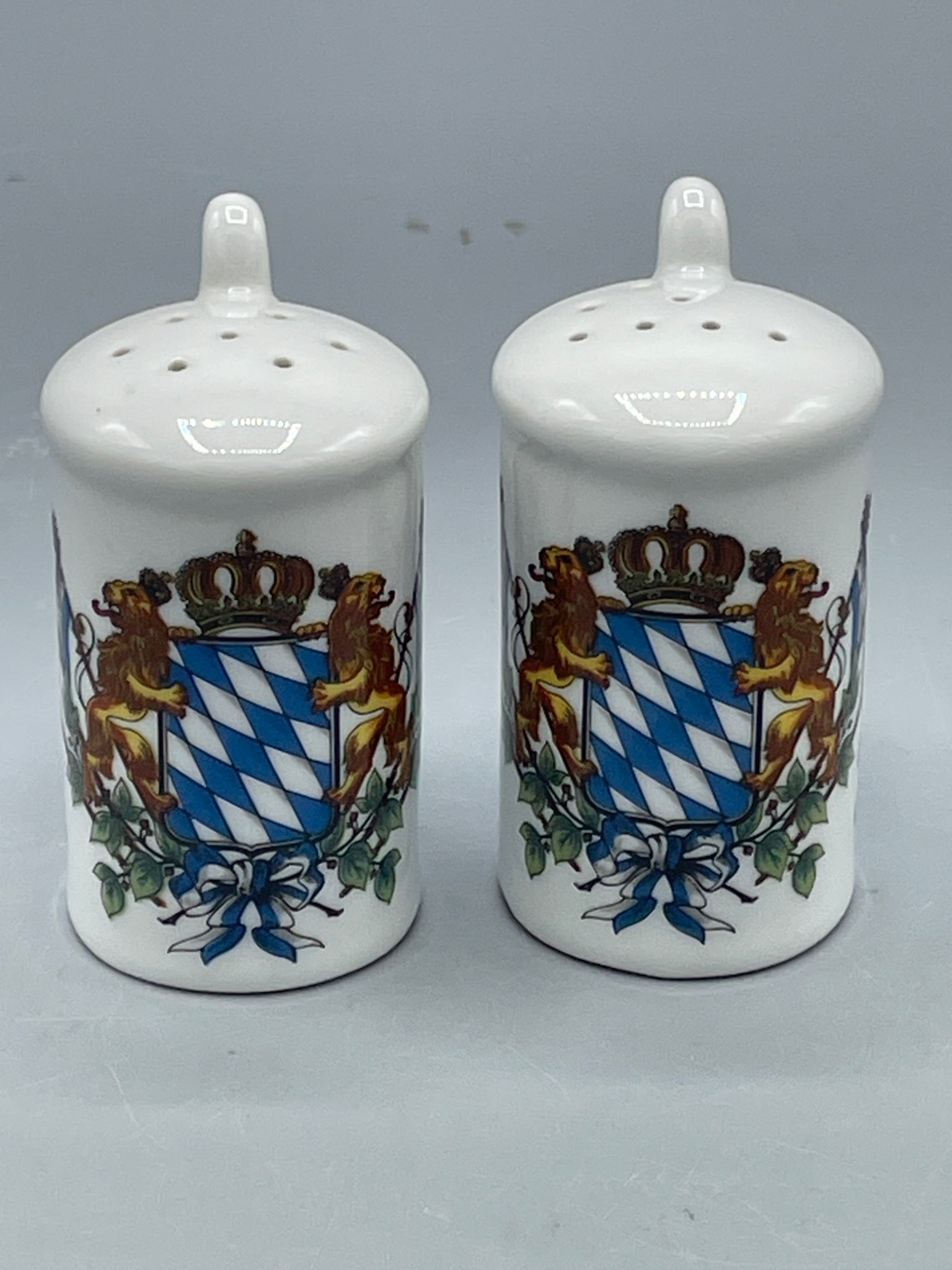 Ceramic Salt and Pepper Shakers with Bavarian Coat of Arms Design: Ceramic salt and pepper shakers featuring the Bavarian coat of arms design. Each shaker includes a handle and multiple perforations on top. Approximately 3 inches in height, as indicated by the ruler