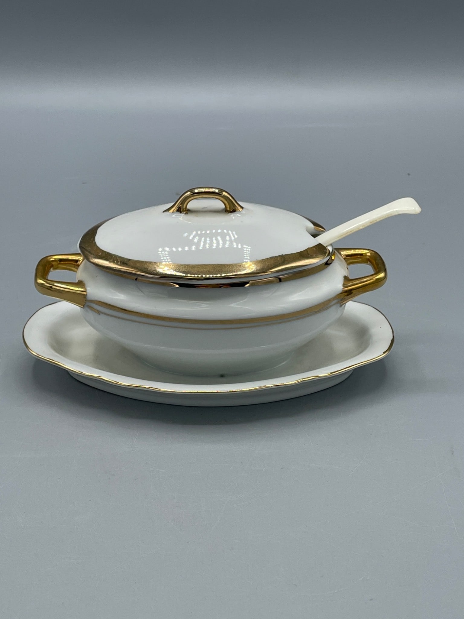 Porcelaine Empire 1282 Covered Serving Dish With Spoon and Tray: This lot includes a Porcelaine Empire 1282 serving dish set, comprising a covered dish, a plate, and a matching spoon. The set features gold-toned accents with a white glaze finish. The base of the