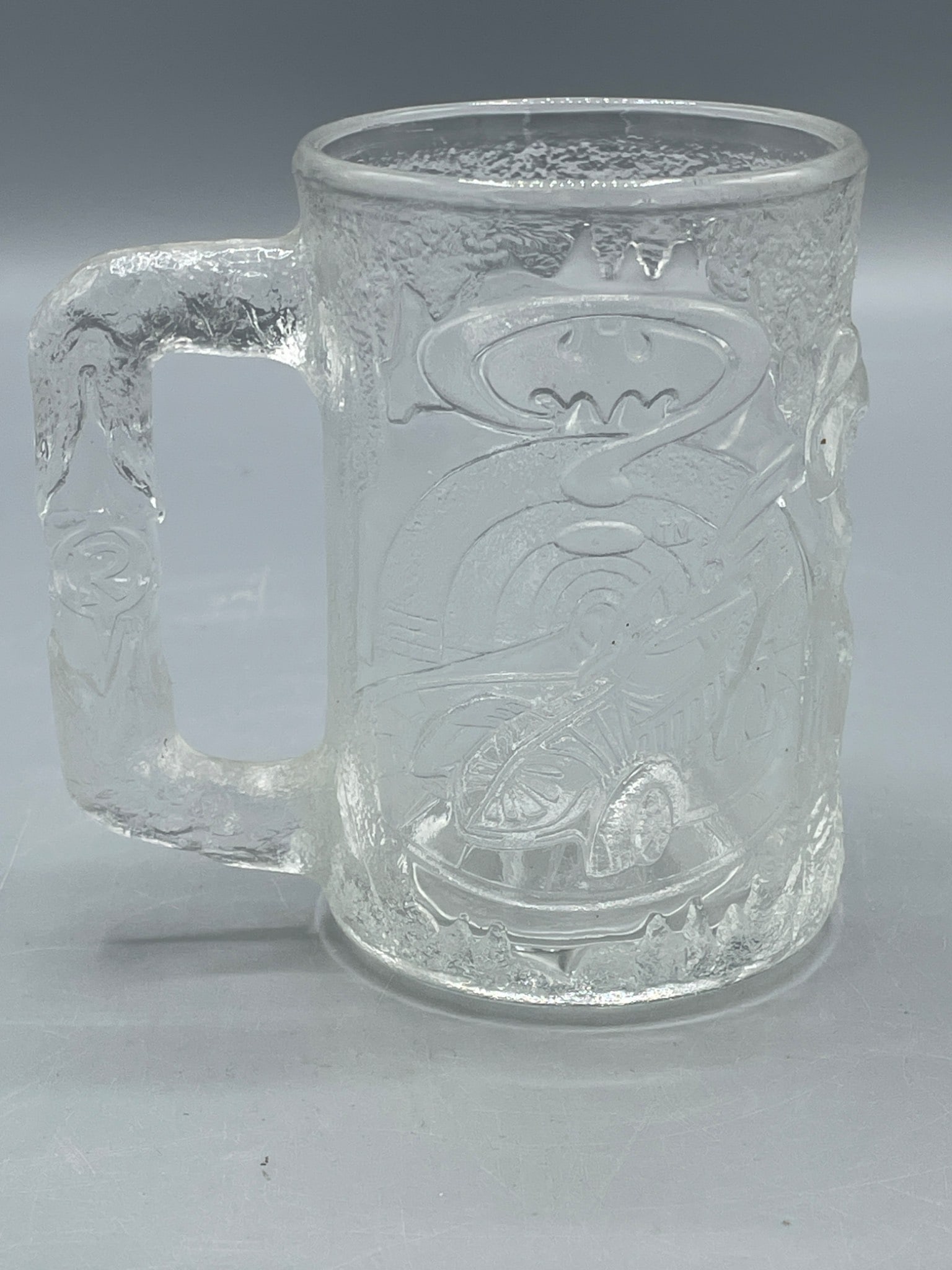 1995 McDonald's Batman Forever Glass Mug Featuring Robin Design: This is a clear glass mug produced by McDonald's in 1995 as part of a promotional series for the movie 'Batman Forever.' The mug features embossed designs of Batman and Robin, with the Batman logo