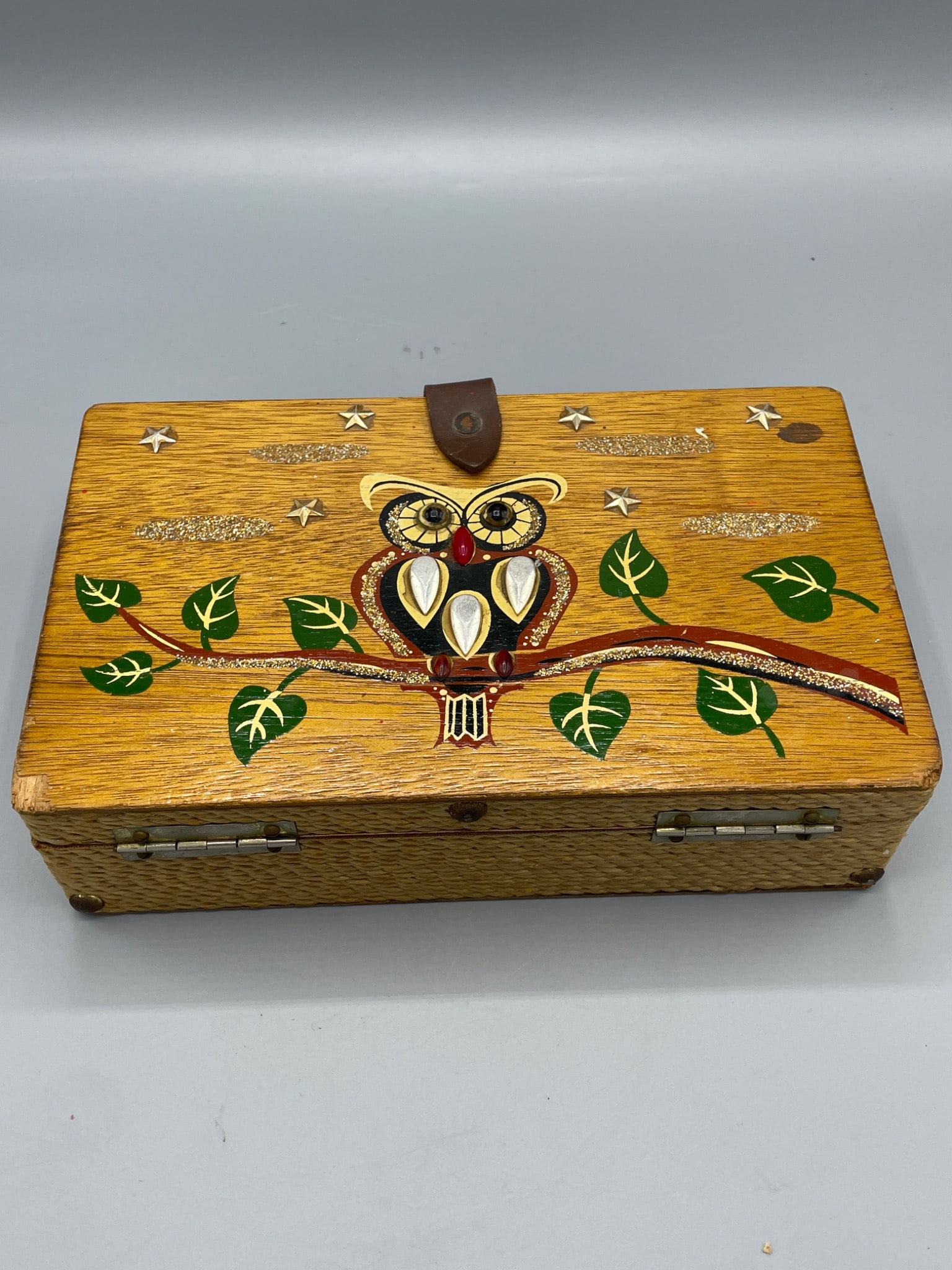 Handmade Wooden Box with Owl Design and Leather Closure: This handmade wooden box features a painted owl design with decorative details such as leaves, stars, and textured accents. It is equipped with a leather strap closure and metal hinges. The box