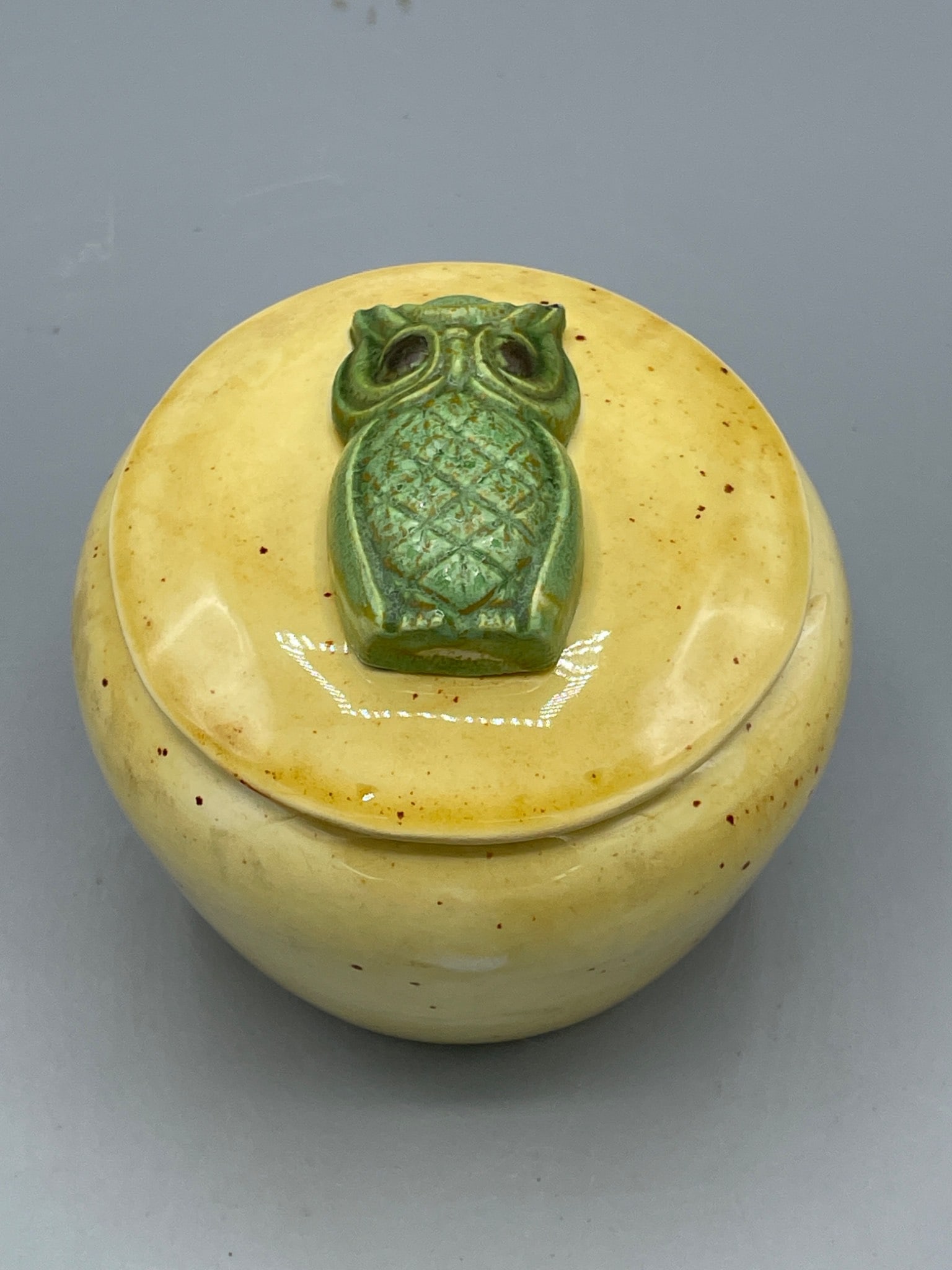 Travis Ceramics Owl-Lidded Ceramic Jar with Yellow Glaze: A small ceramic jar with lid, featuring a green owl-shaped handle. The jar is finished in a yellow glaze with speckled brown details. Produced by Travis Ceramics as indicated on the base stamp and