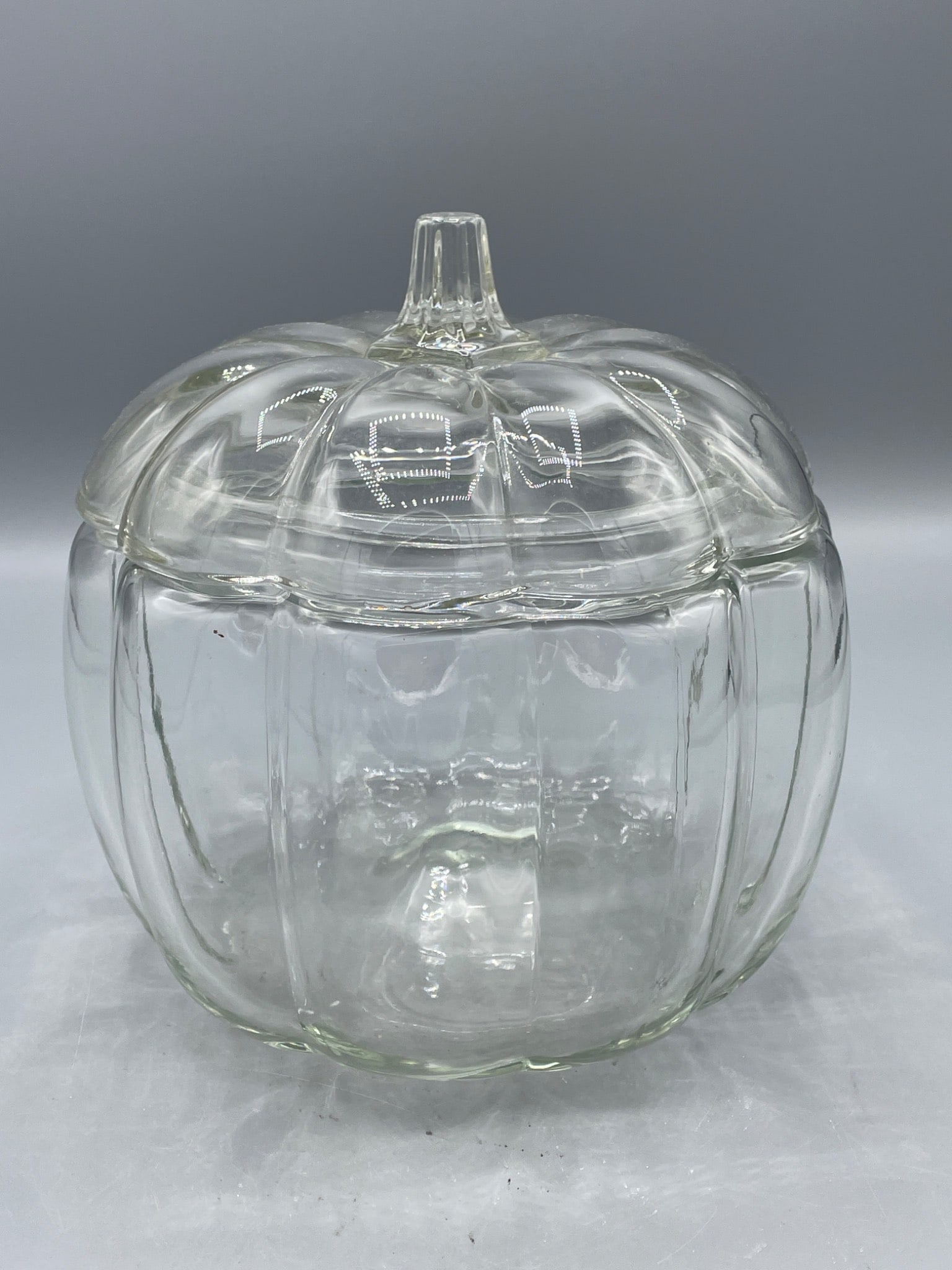 Glass Pumpkin-Shaped Lidded Container with Clear Finish: A glass pumpkin-shaped container with a lid, entirely clear in finish. The container measures approximately 6 inches in height with the lid and roughly 6 inches in width at its widest point. No