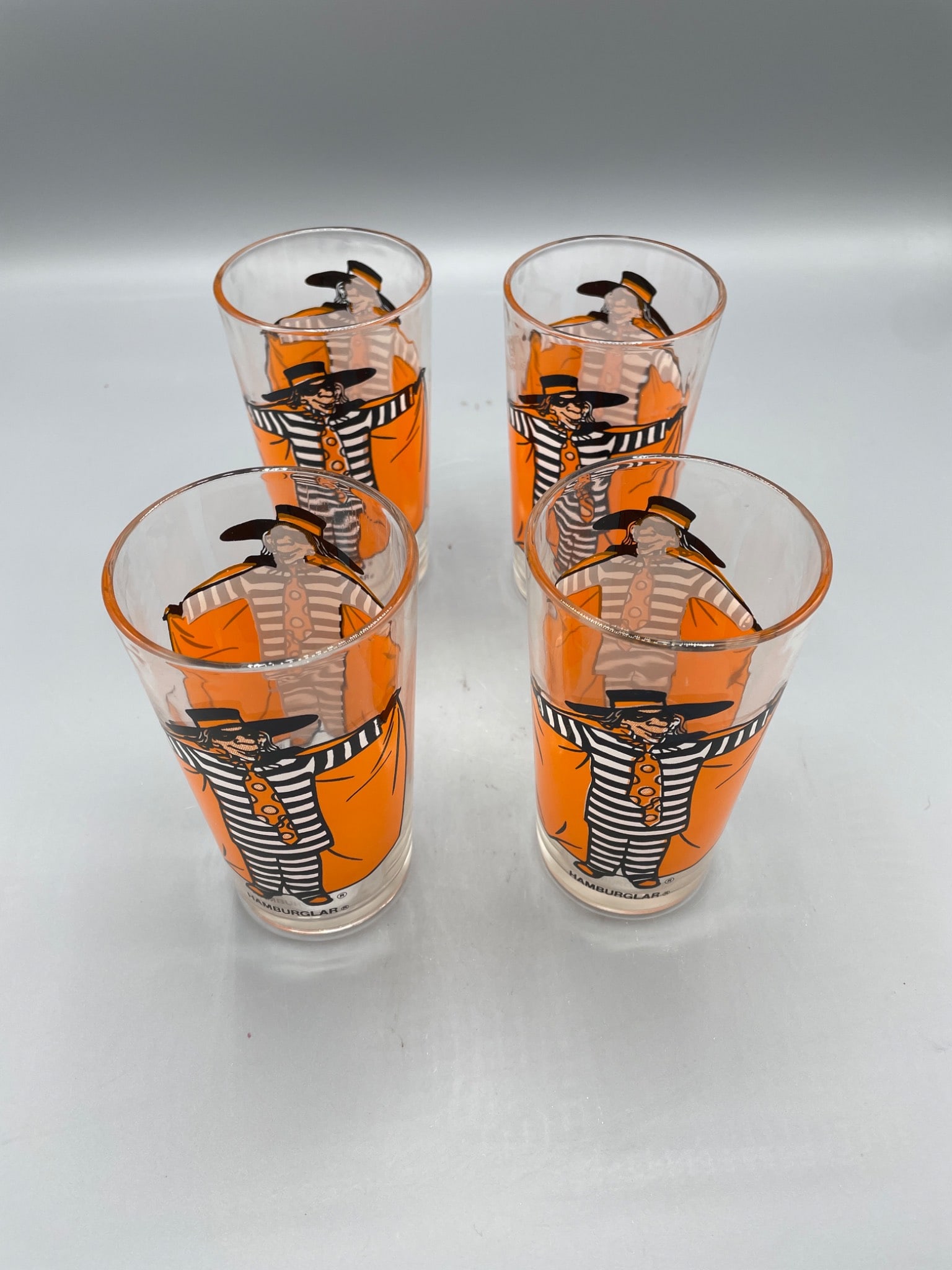 4 McDonald's Hamburglar Character Glasses, Approximately 6 Inches Tall: 4 McDonald's promotional glasses featuring Hamburglar character graphics. Each glass measures approximately 6 inches in height. The designs include vivid orange and black detailing, as well as the