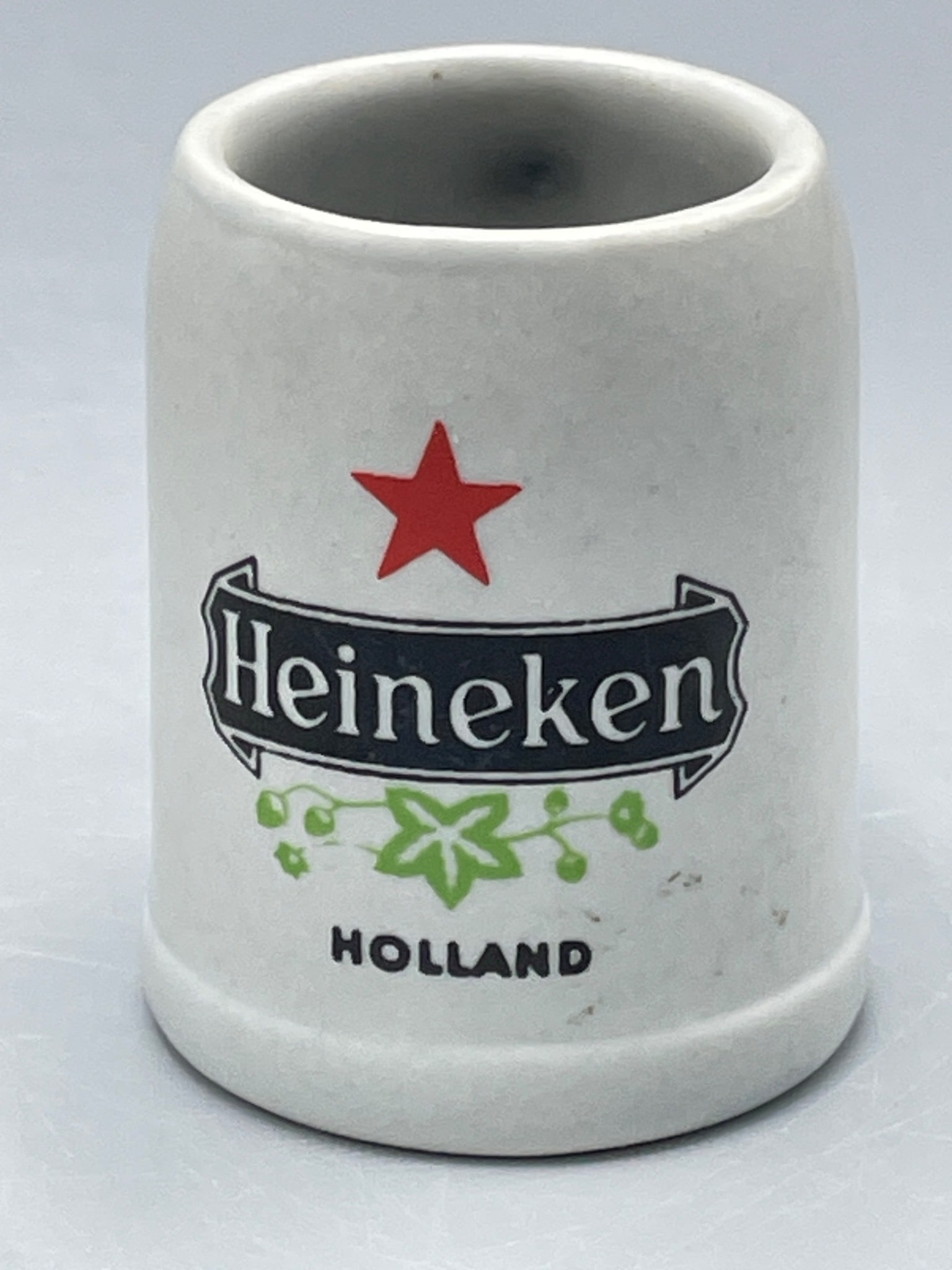 Ceramic Heineken Miniature Mug with Handle by Ceramarte, Brazil: This is a ceramic miniature mug featuring the Heineken logo and 'Holland' text. Manufactured in Brazil by Ceramarte, as identified by the label on the base. The mug is approximately 2.5 inches tall