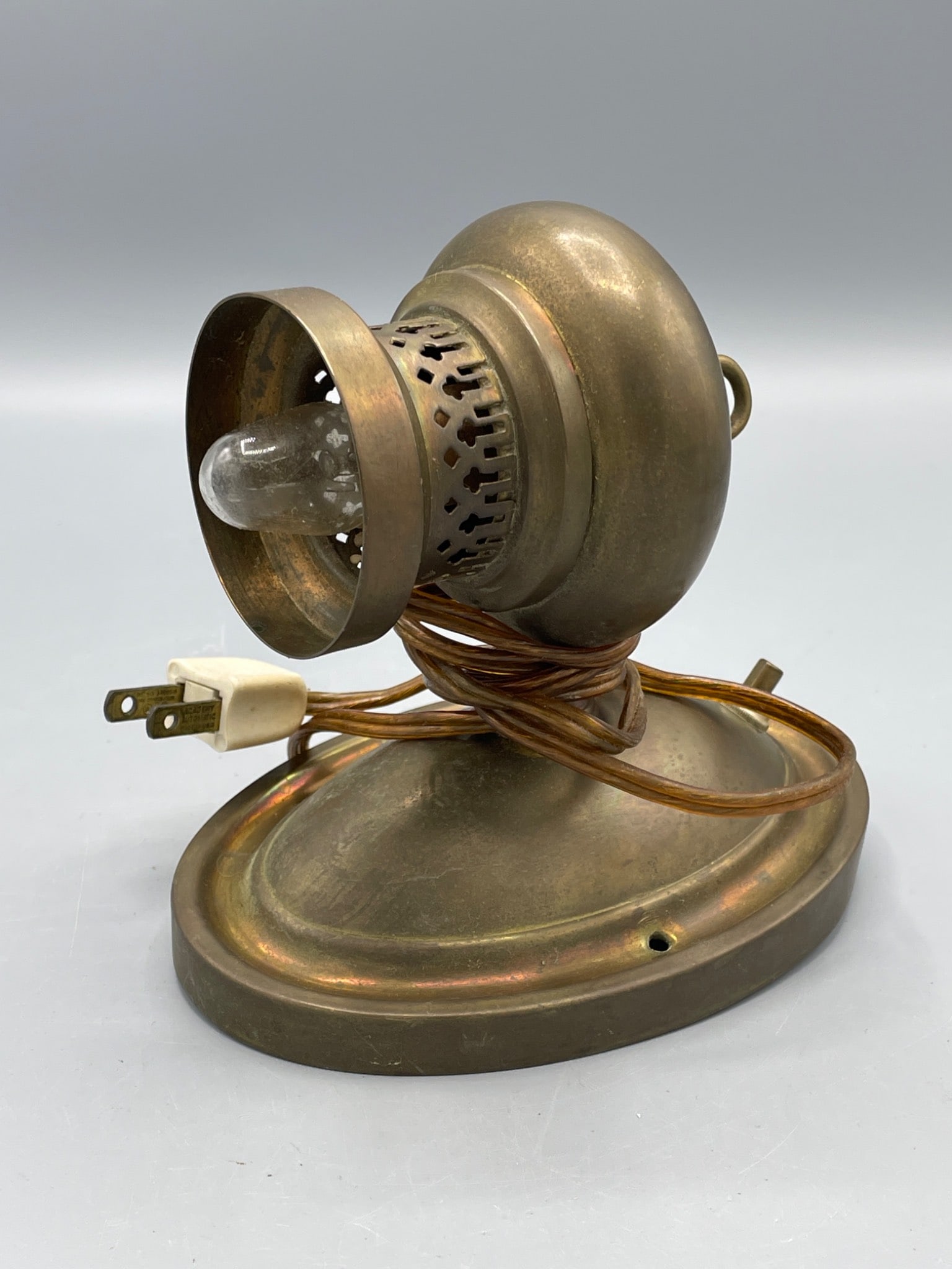 Brass Wall-Mounted Light Fixture With Electrical Wiring: A brass wall-mounted light fixture with perforated detailing around the bulb holder. The fixture has an oval base and includes an attached electrical cord with a two-prong plug and connectors.