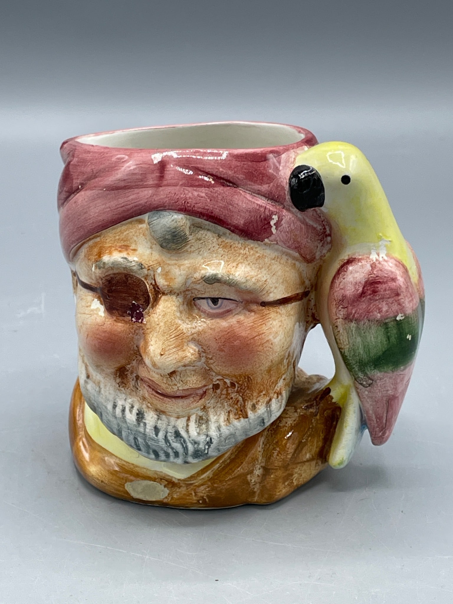 Ceramic Toby Pirate-Themed Mug with Parrot Handle, Approximately 4.5 Inches Tall: This ceramic mug features a pirate face design with a parrot handle. Measuring approximately 4 inches in height, the item shows signs of wear, including paint chipping in some areas. The