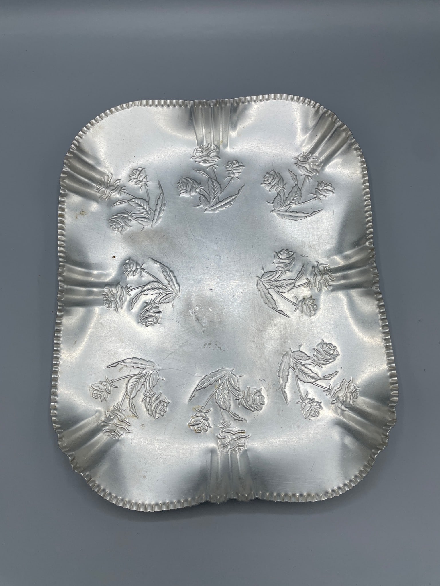 Metal Rectangular Embossed Tray With Floral Motif: This rectangular metal tray features embossed floral patterns on its surface, with scalloped edges and measurements approximately 10 inches in width and 14 inches in length. The tray has an unmarked