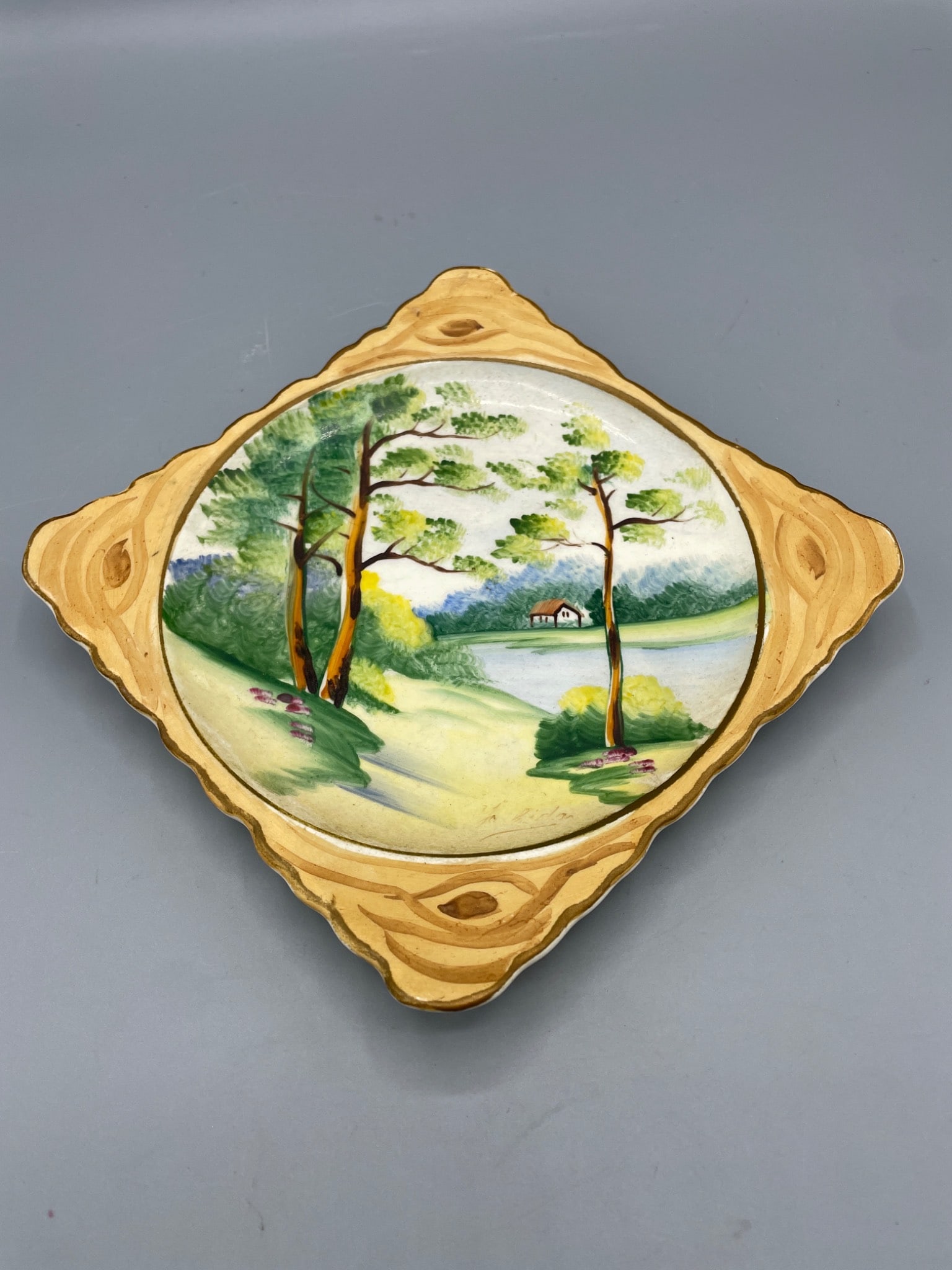 Hand-Painted Square Ceramic Plate with Landscape Design: Square-shaped hand-painted ceramic plate featuring a scenic landscape of trees, a lake, and a house. The outer edges are styled with decorative details. The reverse side bears a small unidentified