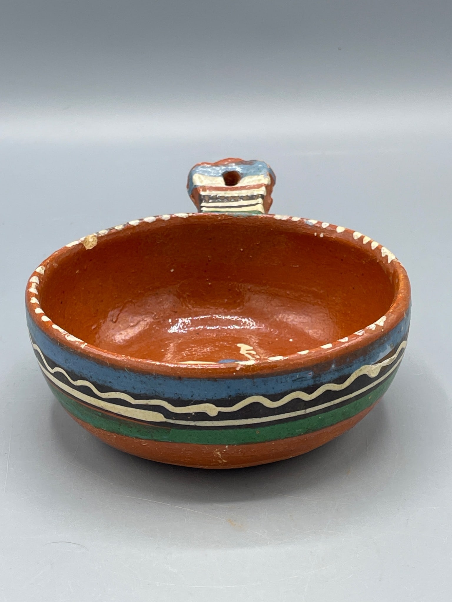 Hand-Painted Ceramic Bowl With Handle and Decorative Pattern: This ceramic bowl features a hand-painted design in multi-colored stripes and abstract patterns. The bowl has a decorative handle with a hole for hanging. It measures approximately 2.5 inches in