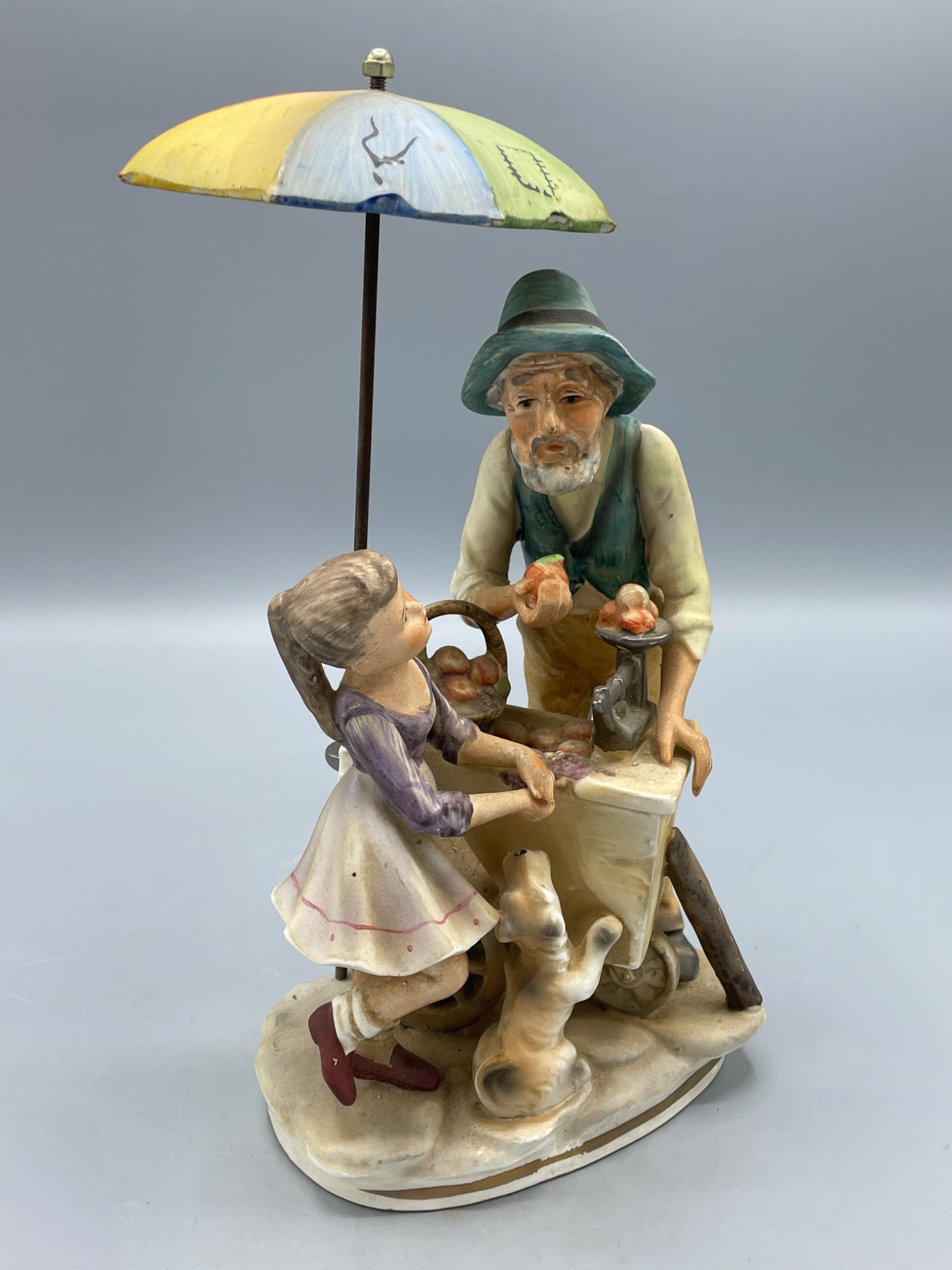 Norleans Japan Ceramic Figurine with Umbrella and Child, Approx. 9 Inches: This is a hand-painted ceramic figurine marked 'Norleans Japan' on the base, featuring an elderly man at a cart interacting with a young girl and a dog. The removable multicolored umbrella adds