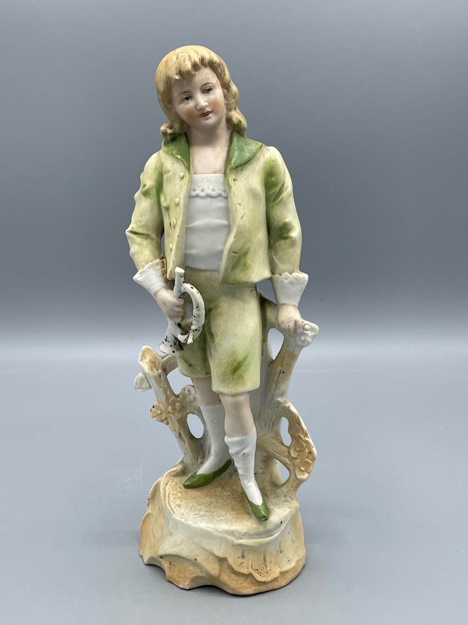 Porcelain Figurine of Boy Holding Horseshoe with Green and Yellow Outfit: This porcelain figurine depicts a boy dressed in a green and yellow outfit, standing and holding a horseshoe. The figurine measures approximately 11 inches in height as shown in the image. The bottom