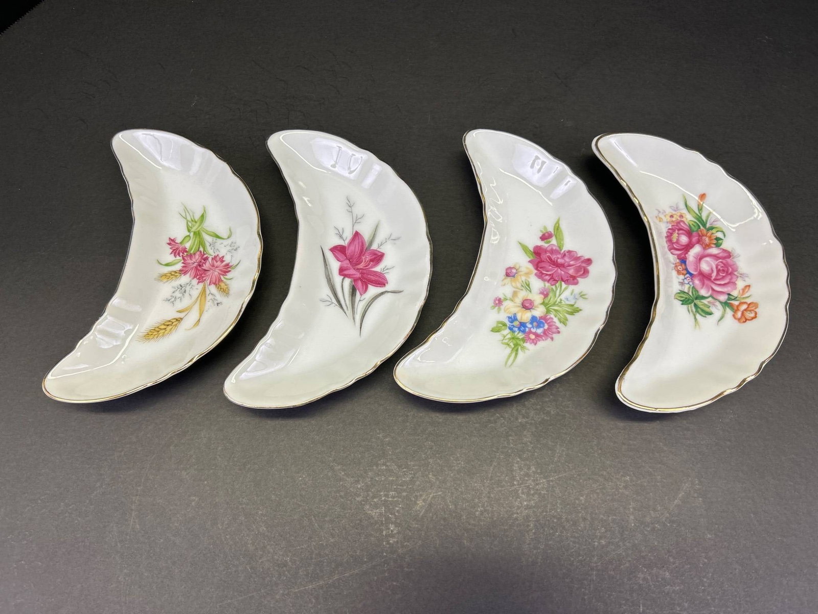 4 Crescent-Shaped Porcelain Dishes with Gold Trim and Floral Motifs: 4 crescent-shaped porcelain dishes, each featuring gold trim and unique hand-painted floral motifs. The back of each dish displays a gold shield logo with a crown and letter 'M', indicating the