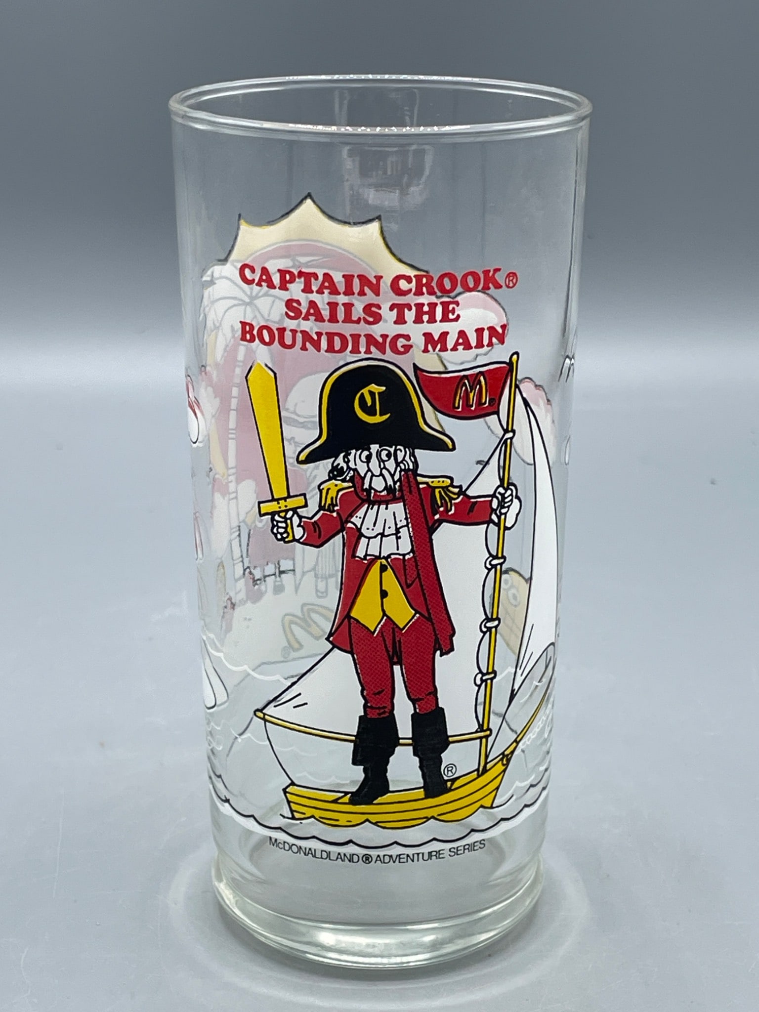 1980 McDonaldâ€™s Captain Crook Glass From McDonaldland Adventure Series: A clear glass featuring Captain Crook from the McDonaldland Adventure Series, produced in 1980 by McDonaldâ€™s Corporation. The design includes Captain Crook on a ship and an island