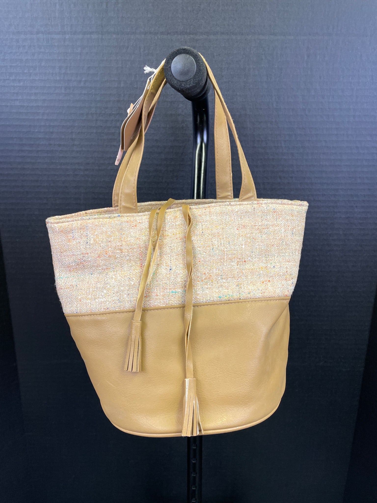 Tan Tote Bag With Woven Top and Leather Bottom: Tan tote bag featuring a woven textile upper portion and a smooth leather bottom. Includes dual leather straps and a drawstring closure with tassels. The bag has a rounded leather base and a fabric