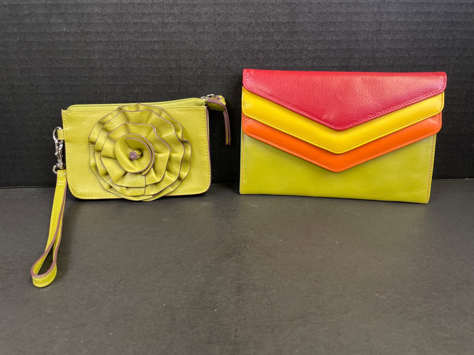 Two Leather Wallets: Green Wristlet and Tri-Color Envelope Clutch: This lot contains two leather wallets. The first is a green wristlet with a floral accent, zipper closure, and multiple card slots inside. The second is a tri-color envelope-style clutch in red,