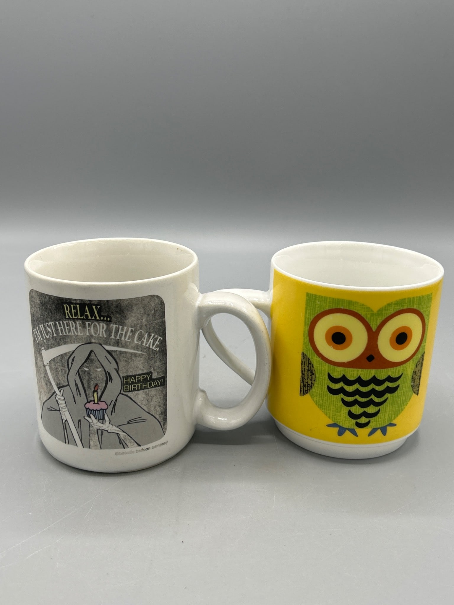 Ceramic Mugs Featuring Grim Reaper and Owl Designs: This lot includes two ceramic mugs: one featuring a Grim Reaper with the text 'RELAX... I'M JUST HERE FOR THE CAKE. HAPPY BIRTHDAY!' and the other with a colorful owl design. Both mugs are marked as