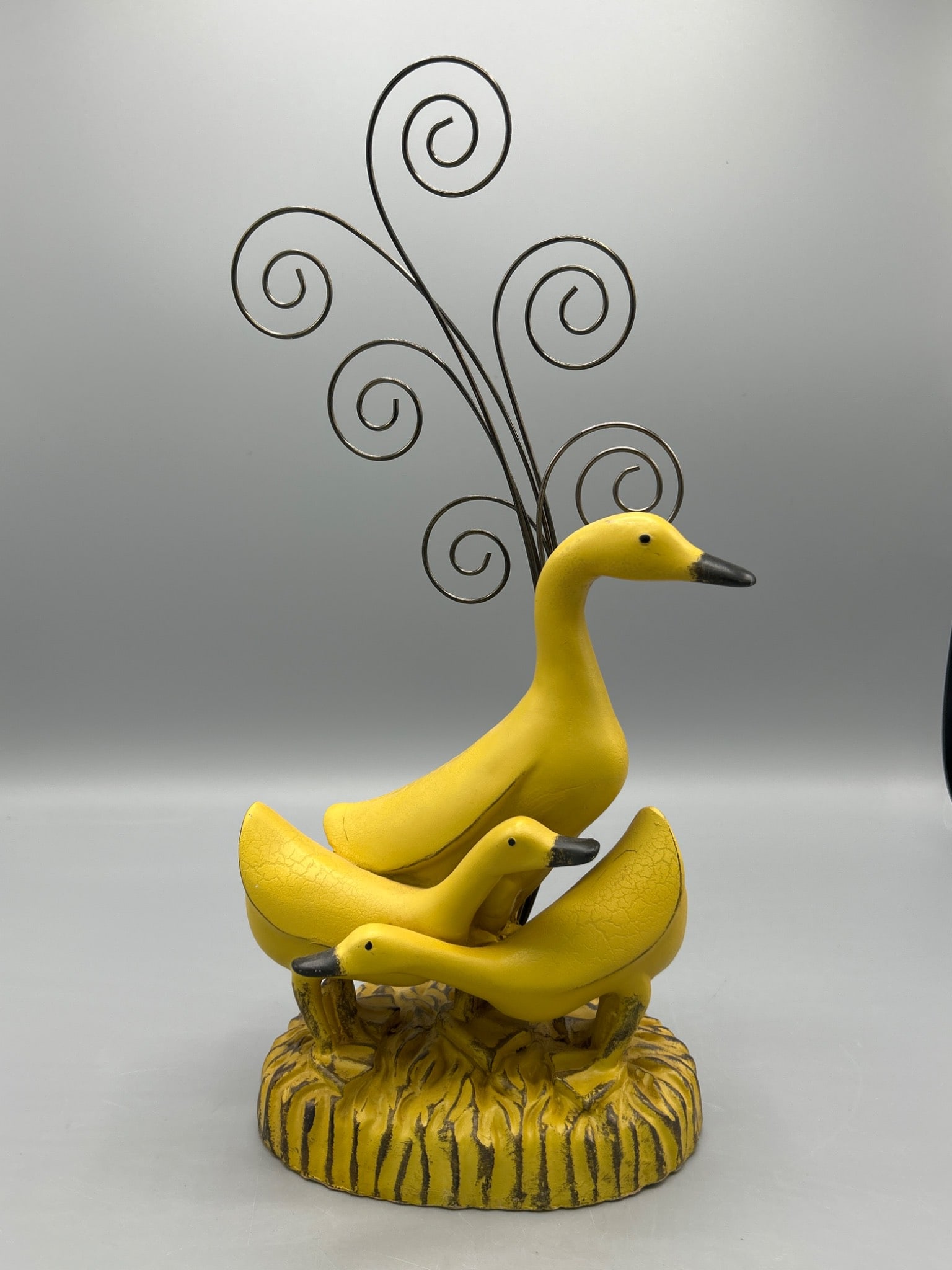 Yellow Duck Family Figurine With Metal Swirls On Decorative Base: Three-piece yellow duck figurine set mounted on a painted decorative base. Includes a tall metal swirl design extending from the base as an accent. Approximate height is 14 inches, including the