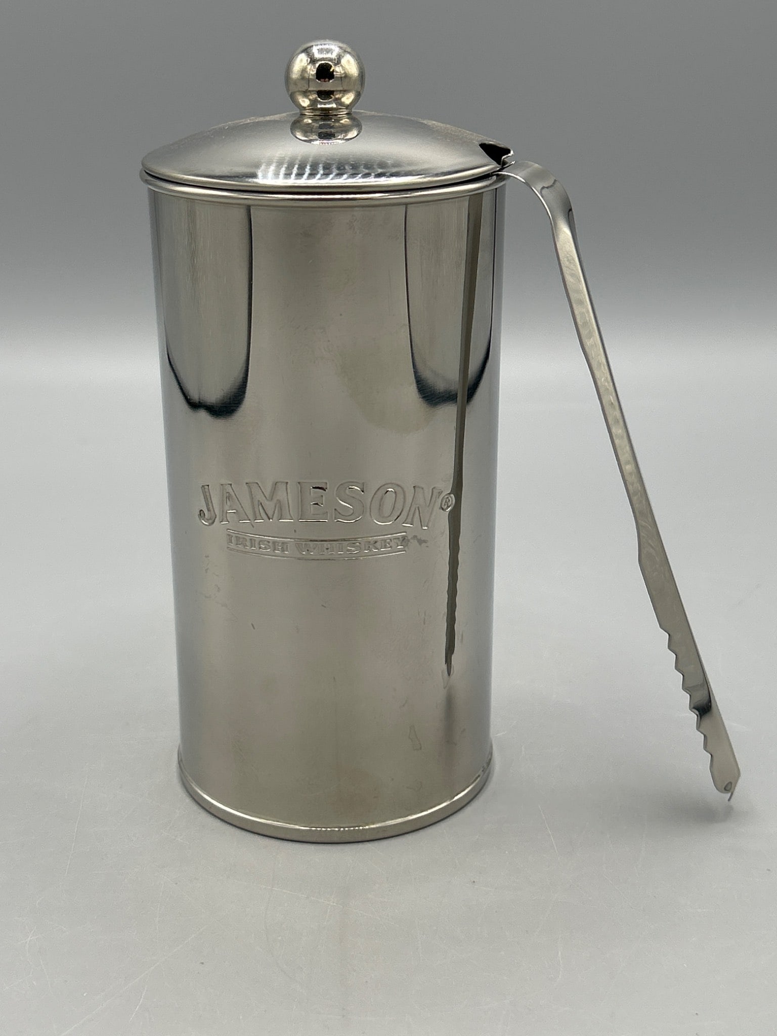 Jameson Irish Whiskey Branded Ice Bucket With Lid and Tongs: Jameson Irish Whiskey branded ice bucket made of stainless steel. Includes lid with decorative knob and matching stainless steel tongs. Identifying label recommends cleaning with warm soapy water only