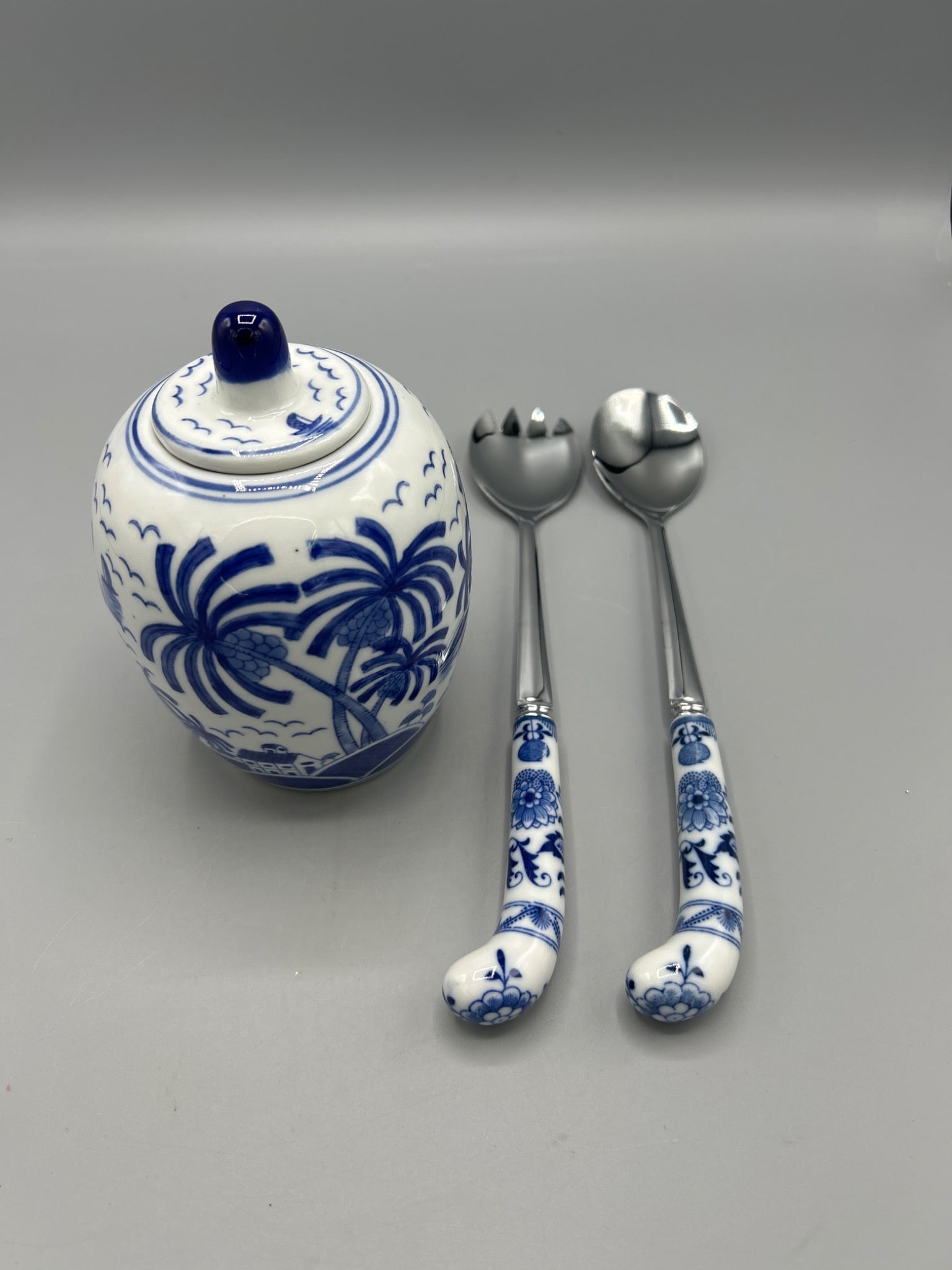 Blue and White Ceramic Jar with Lid and Matching Salad Servers: This lot includes a blue and white ceramic jar with lid featuring a palm tree motif, accompanied by a matching pair of salad servers. The jar measures approximately 6 inches in height, while the