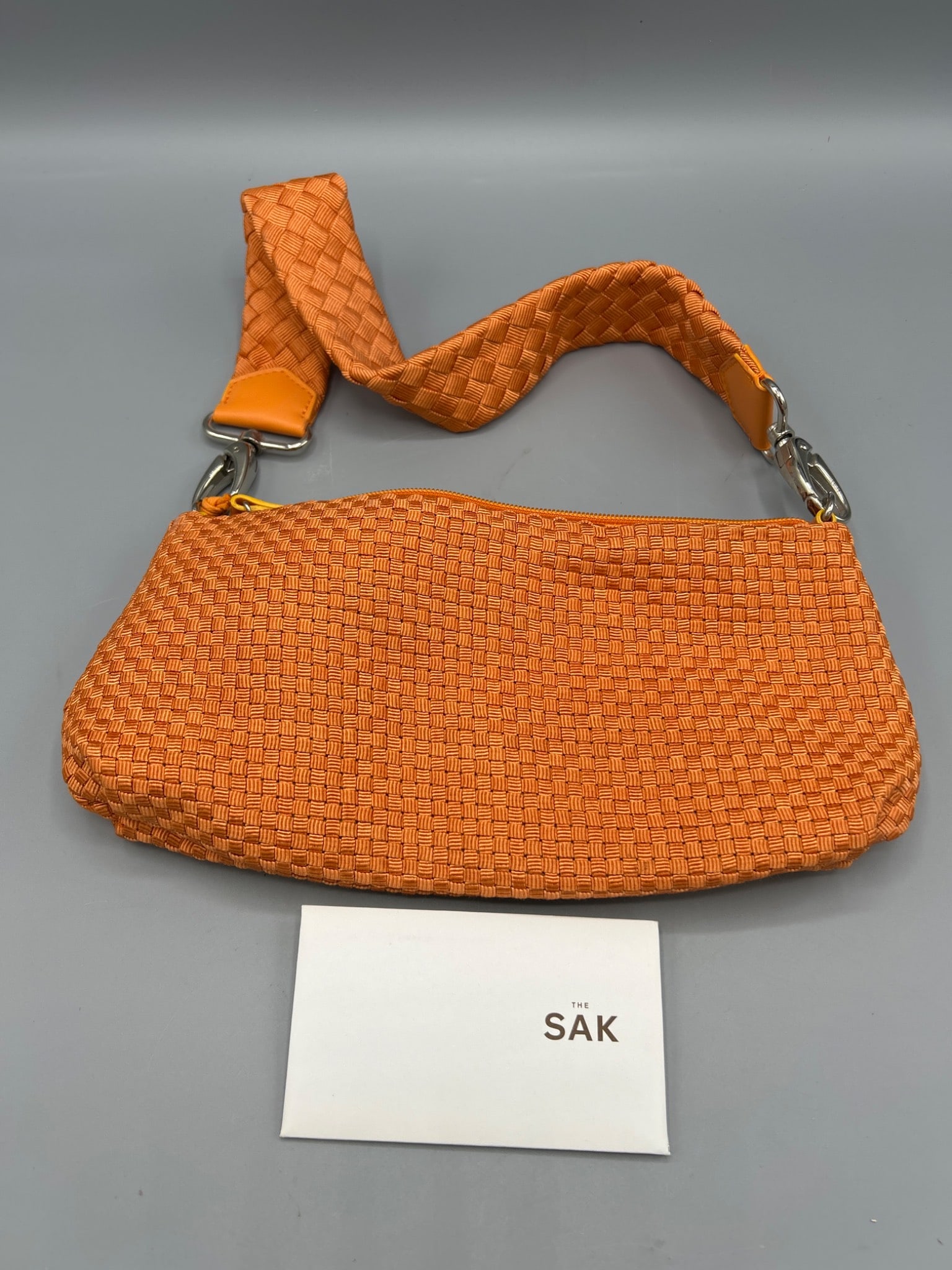 The Sak Orange Woven Handbag with Adjustable Strap: This is an orange woven handbag from The Sak, featuring an adjustable fabric strap with leather accents. It has an orange zip closure and a metal branded decoration. The inner lining is orange and