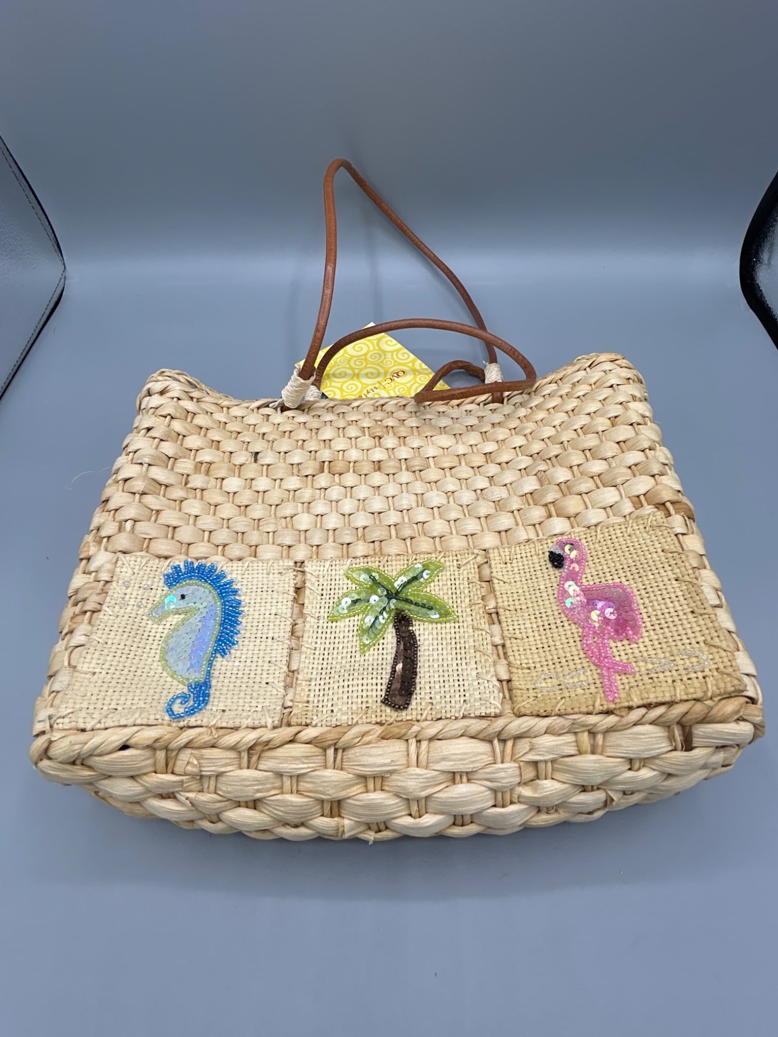 Woven Straw Tote Bag with Embroidered Design and QVC Tag: This woven straw tote bag features decorative embroidered panels including a seahorse, palm tree, and flamingo. The interior is lined with a blue fabric featuring a dotted pattern, secured by a