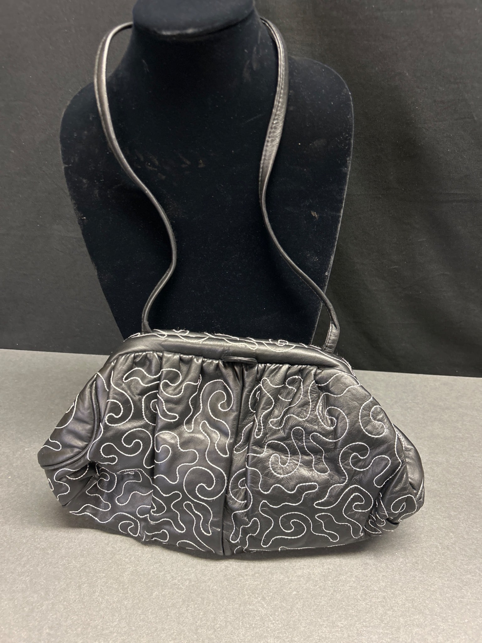 Black Leather Purse With Embroidered White Swirl Design: This black leather purse features an embroidered white swirl pattern. It has a single compartment with a zippered interior pocket and a snap closure. The bag includes a shoulder strap, as shown. The