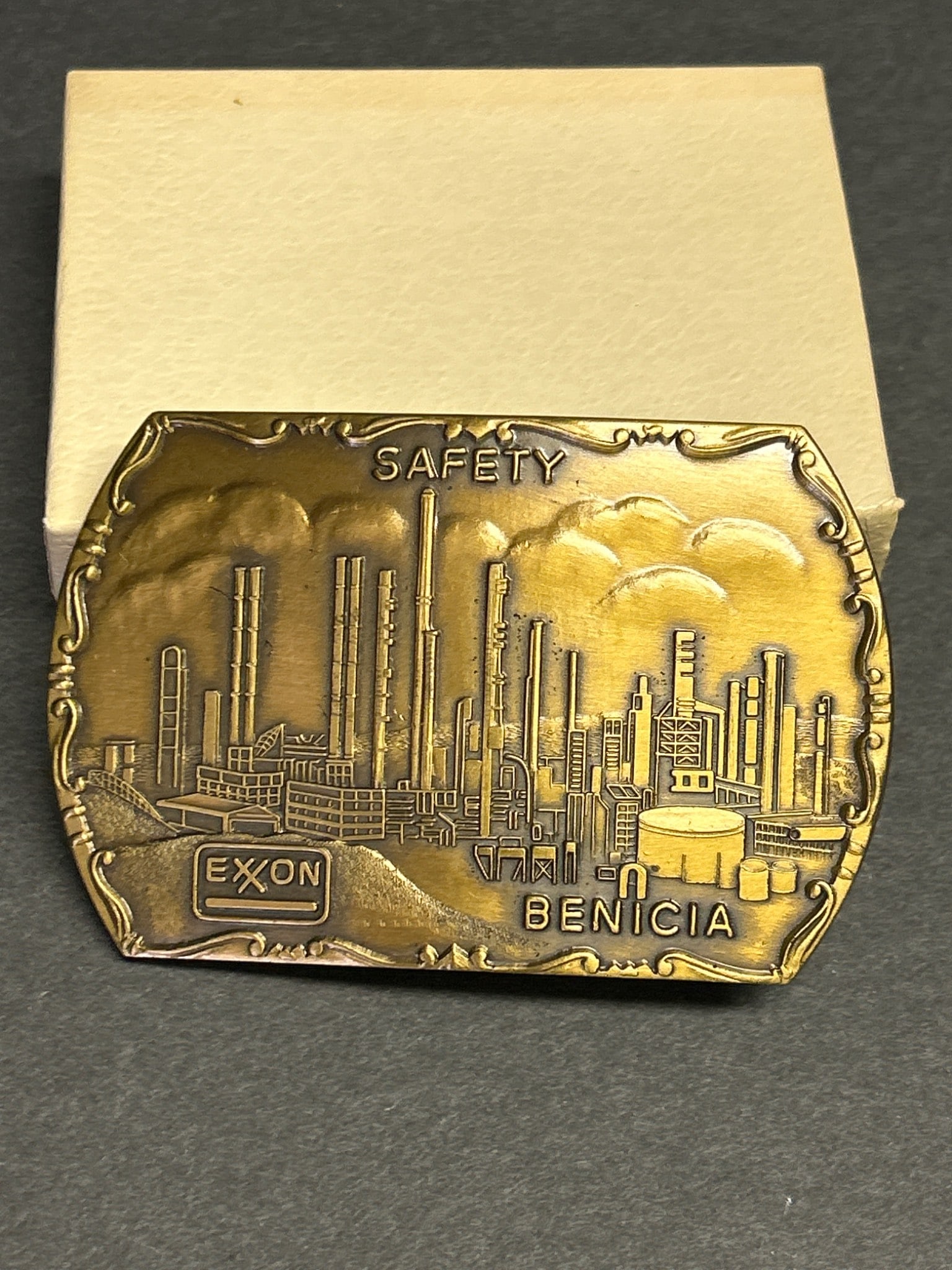 Brass Belt Buckle Featuring 'Exxon Safety Benicia' Design: This brass belt buckle depicts a detailed design of an industrial refinery scene with the text 'Exxon Safety Benicia.' It appears to be cast in solid brass and includes attachment hardware on the