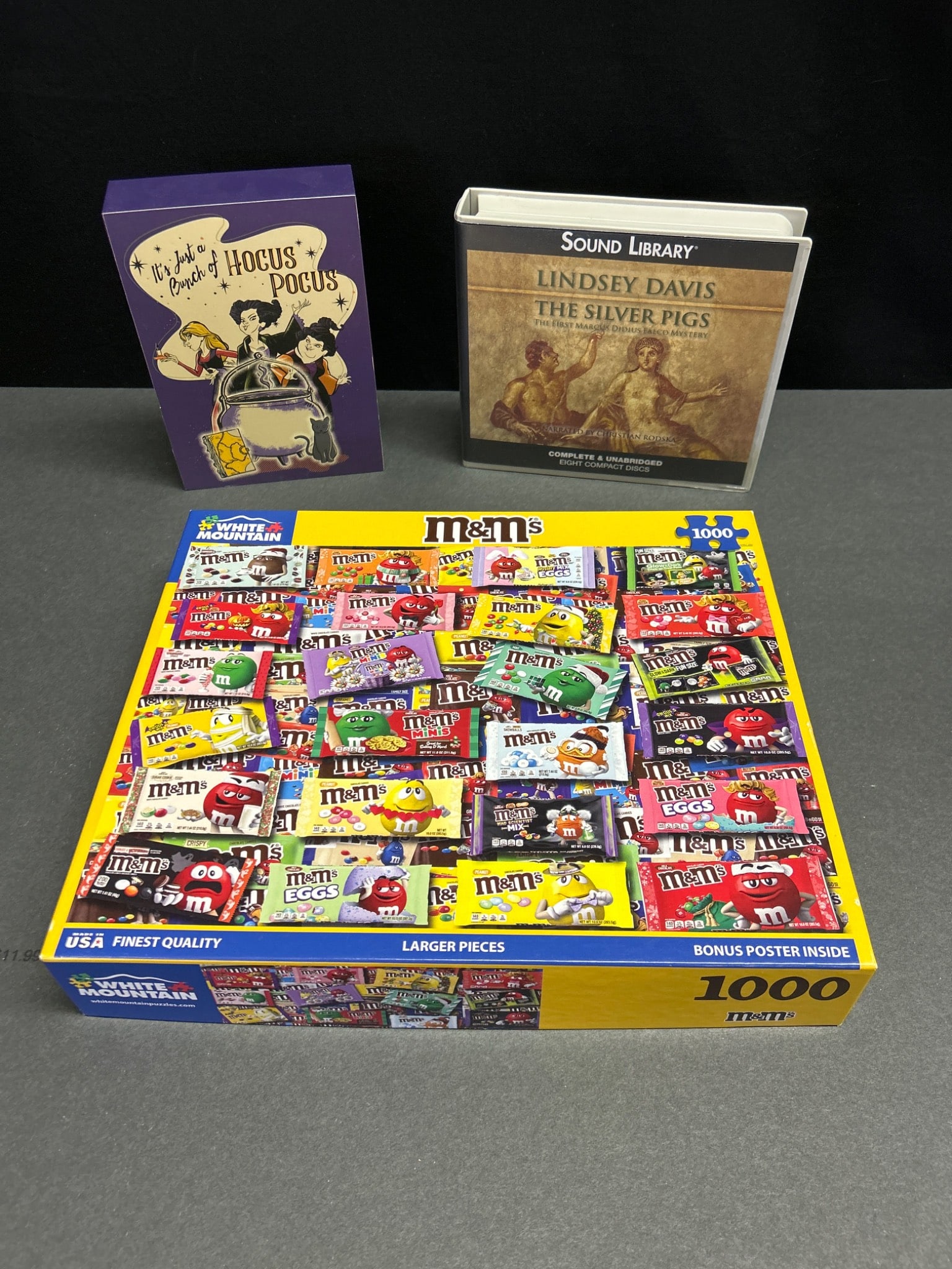 White Mountain M&M's Puzzle, Lindsey Davis Audiobook, and Hocus Pocus Deck: This lot includes a 1000-piece White Mountain 'M&M's' puzzle, model 172-GR1, a complete and unabridged Sound Library audiobook of Lindsey Davis' 'The Silver Pigs' on eight compact discs, and a box set
