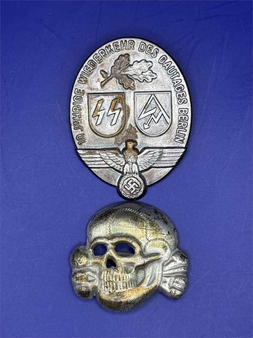 WW2 German Oval Badge and Skull pin: Dimensions: 1.5in and 1inThe image displays two pieces of German World War II (WWII) militaria: a commemorative SS/SA tinnie badge and a Totenkopf (Death's Head) insignia. The oval badge is a commemor