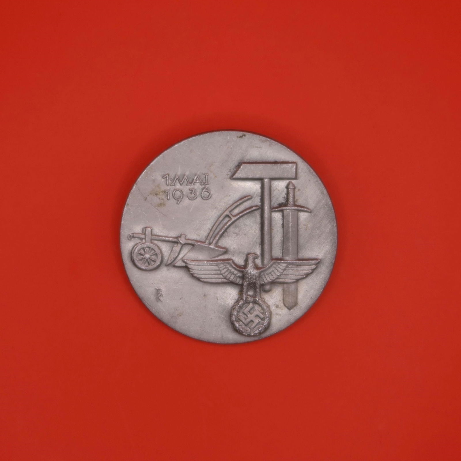 1936 Tag der Arbeit Tin Day Badge: This round 1936 German "Tag der Arbeit" badge is made of a light-colored metal, likely tin. The front shows a plow, hammer, and sword grouped together above an eagle emblem. The date "1. Mai 1936" app