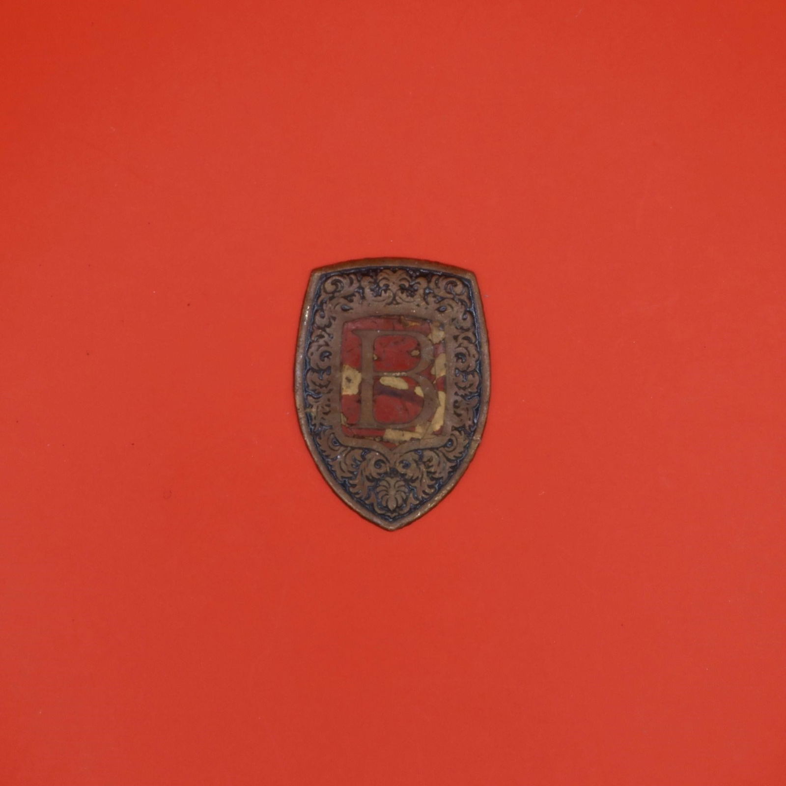 German Metal Shield Badge "B": This is a small metal shield badge with ornate scrollwork and a prominent red "B" at the center. The design suggests it was a decorative or uniform-style emblem, likely representing an organization, s