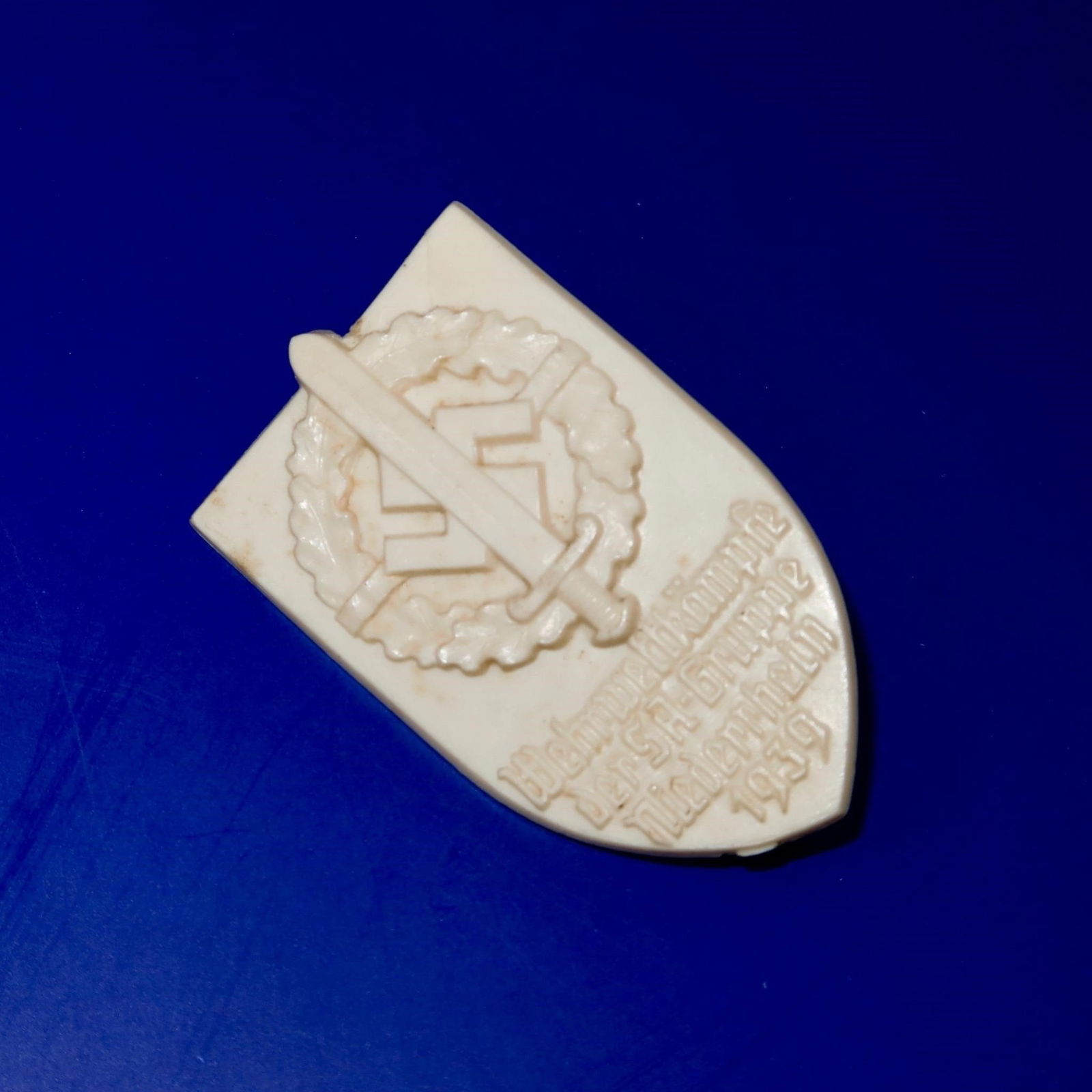 1939 SA Group Niederrhein Sport Day Badge: White protoplastic badge, approximately 35 × 50 mm, originally with bronze-colored paint that has faded over time. Features a sword with SA insignia on the pommel over an oak leaf wreath, with