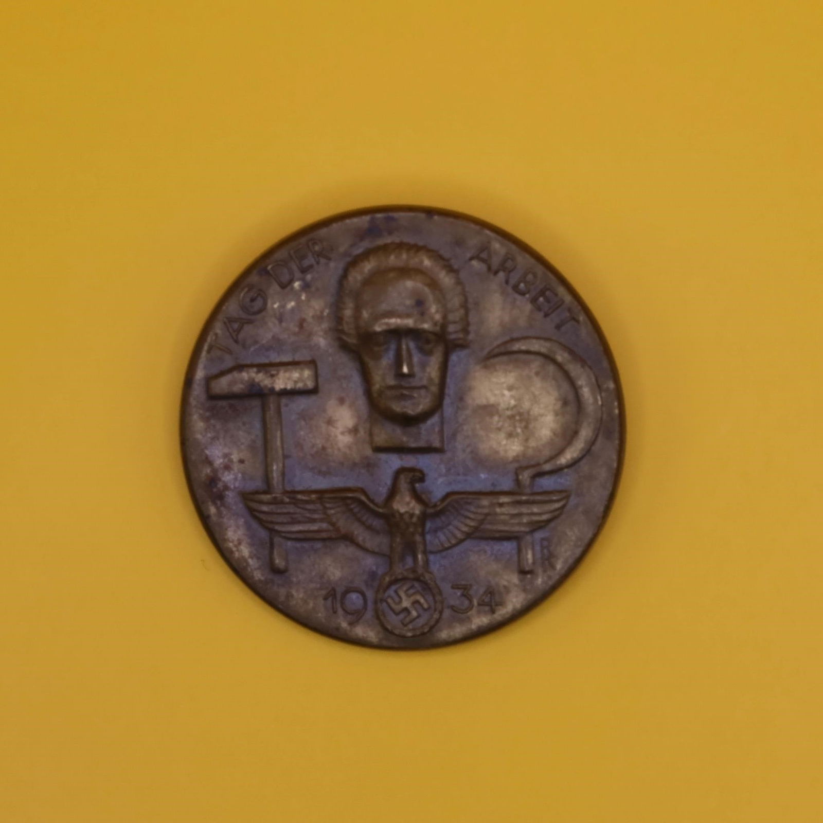 1934 German Labor Day Badge (Bronze Tone): A round 1934 commemorative German Labor Day badge in bronze tone. The front shows a hammer, a sickle, and a stylized face above an eagle emblem. The back has the original pin, though aged. Displays