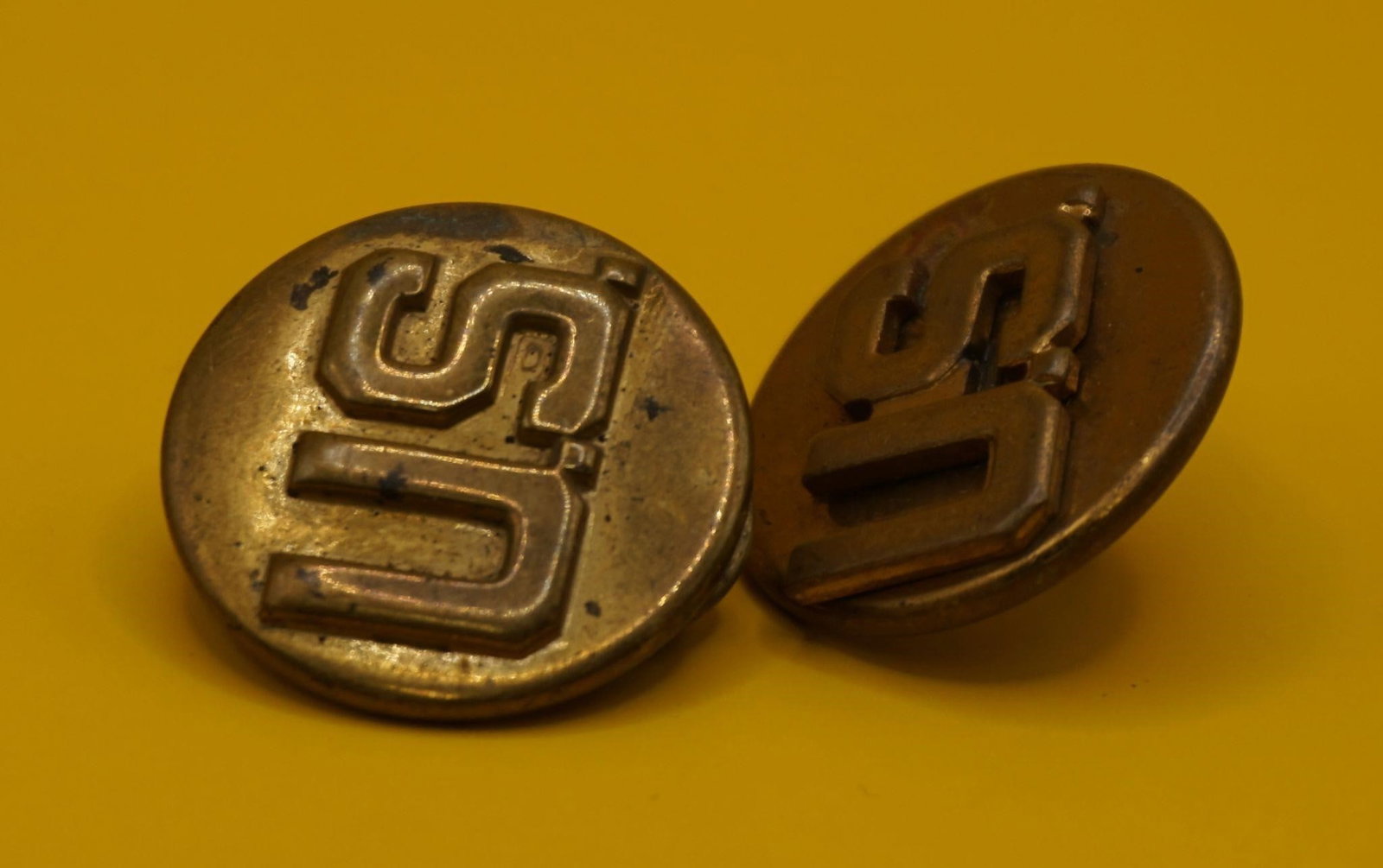 WWII Vintage US Military Brass Collar Insignia Pin: These are vintage US military brass collar insignia pins. They feature the letters "US" and were typically worn on the collar of a dress uniform. The pins are made of brass and show signs of tarnish