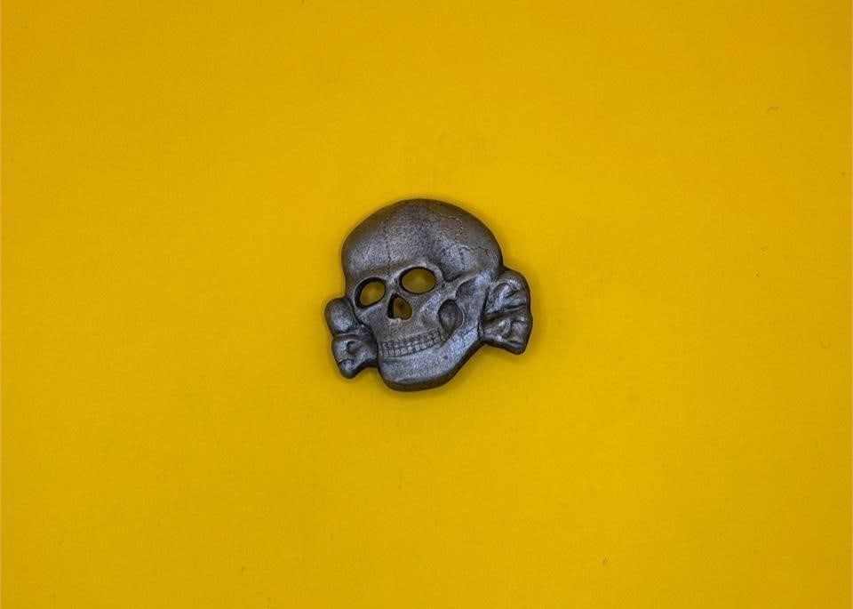 Late War Assmann SS Skull: The SS skull (Totenkopf) was worn on SS caps from 1934 through 1945, symbolizing membership in the Schutzstaffel. Late-war Assmann examples were die-stamped in zinc with a silver wash, reflecting