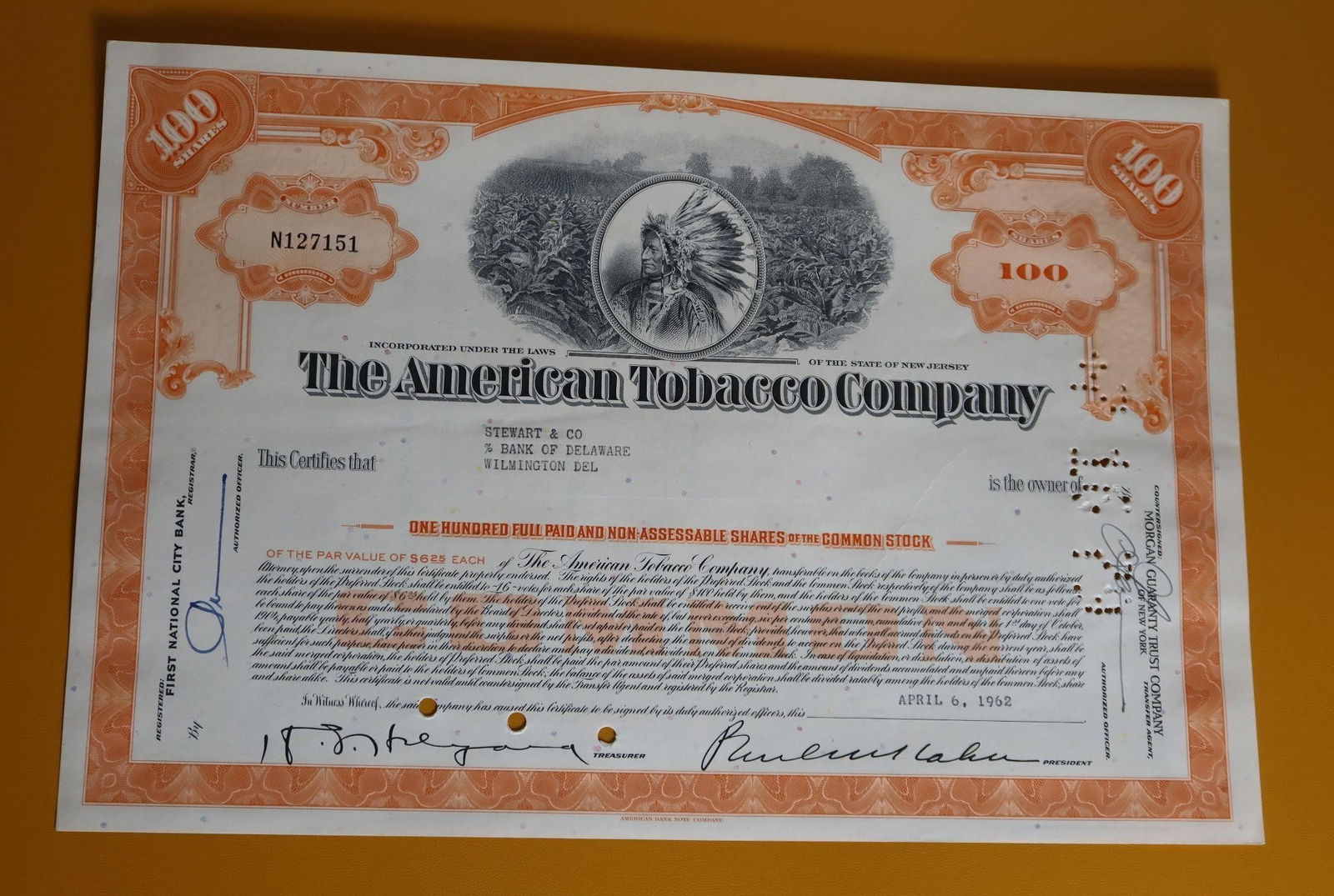 American Tobacco Company Stock Certificate: A vintage American Tobacco Company stock certificate issued on April 6, 1962. The certificate is for 100 shares of common stock. It features a vignette of an American Indian chief, often identified as